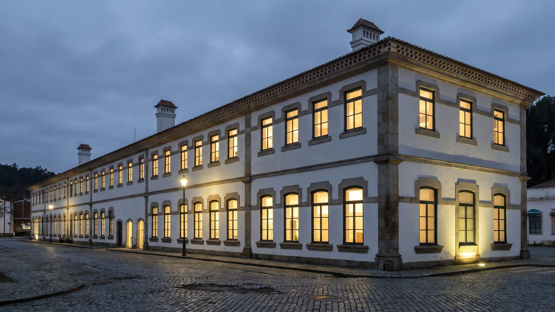 The long facade of the Caldas da Felgueira bath house at dusk, its windows lit above wet cobbles