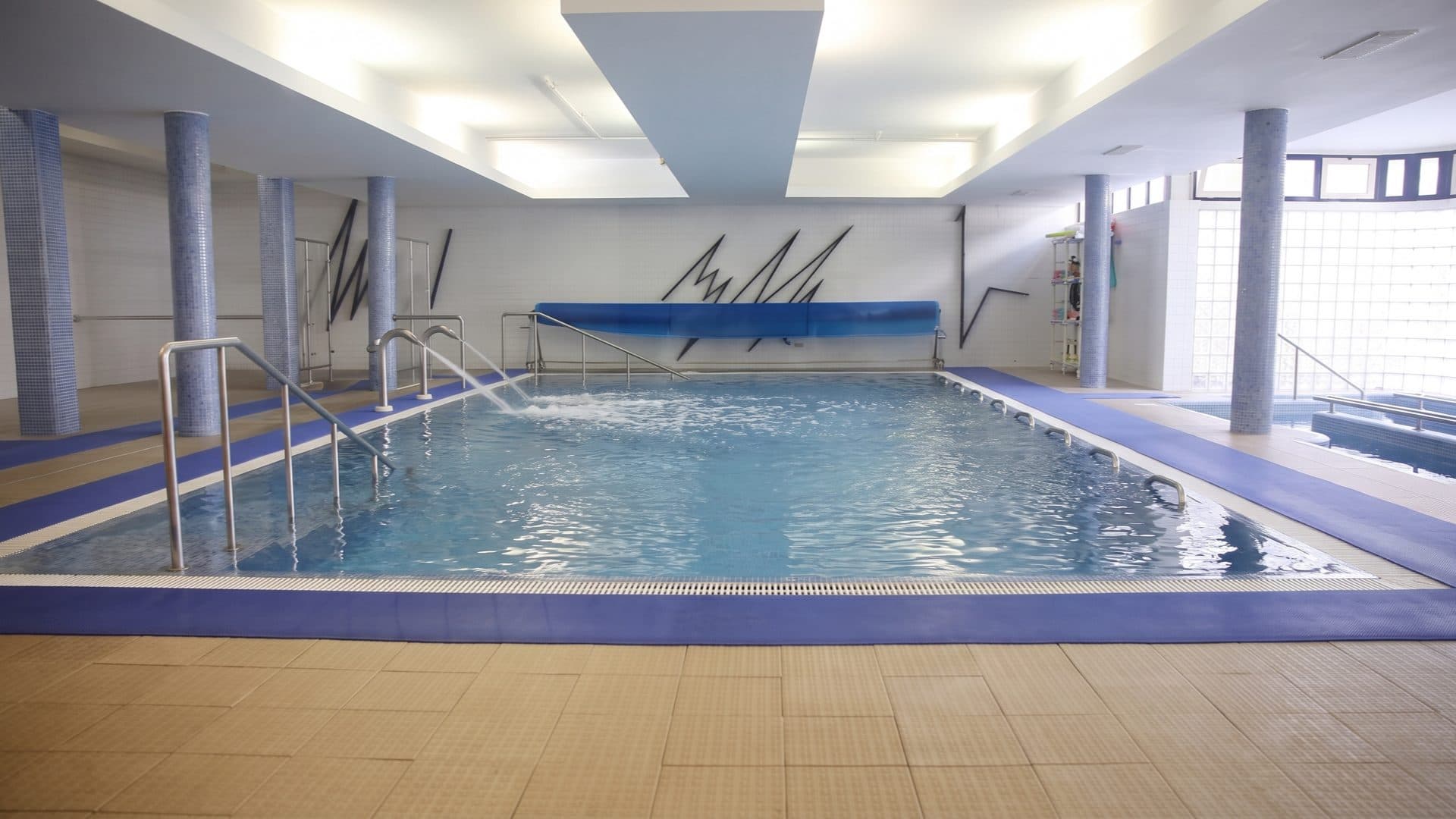 Indoor therapy pool with a water jet playing across the surface, blue-mosaic columns and glass-block windows.