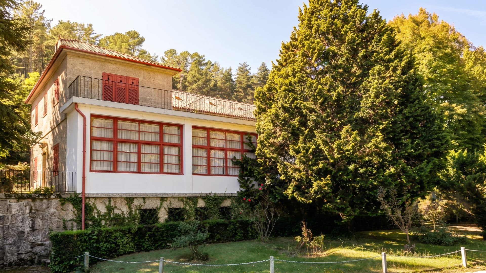 Historic spa villa with a red-framed glass veranda and red shutters under a tiled roof, set among pines and a tall cypress on a lawned slope.
