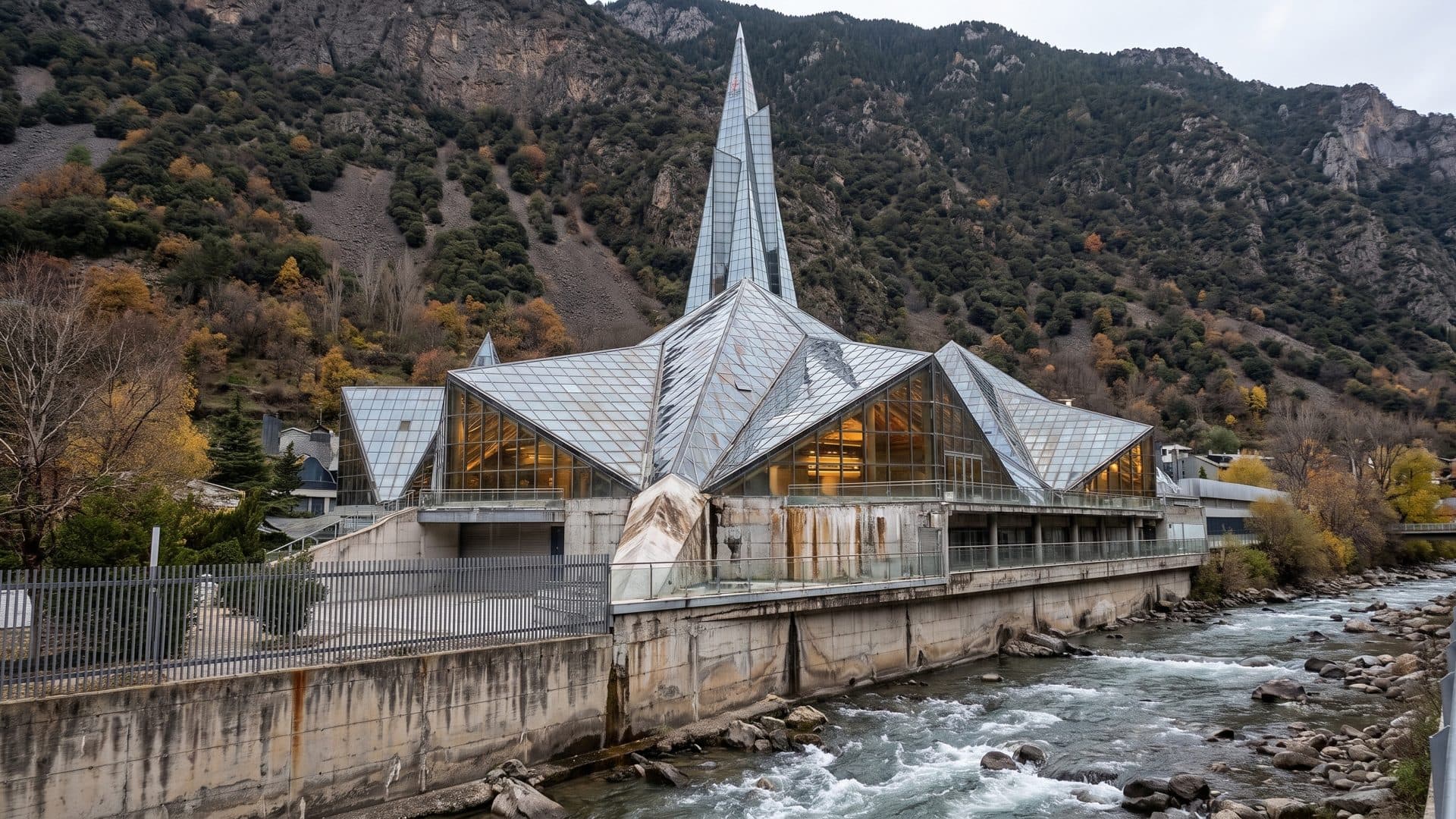 The glass spire and glazed pavilion of Caldea rising above the mountain river at Escaldes-Engordany, with the wooded valley slope behind