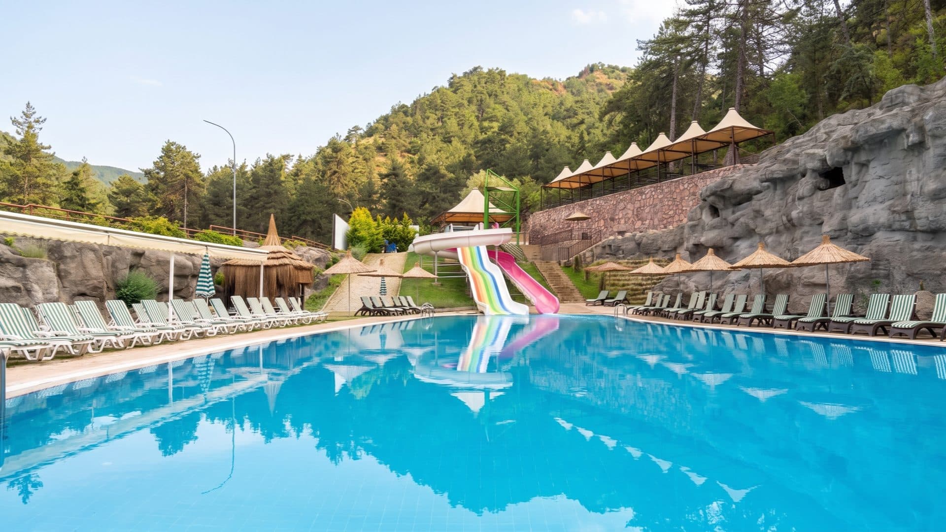 Large outdoor thermal pool ringed by straw parasols and loungers, with rainbow and pink water slides and a pine-clad hillside behind.