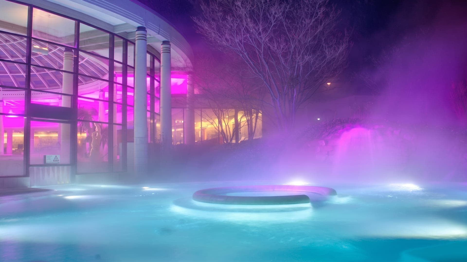 Outdoor pool lit magenta and blue at dusk, with steam rising from the water