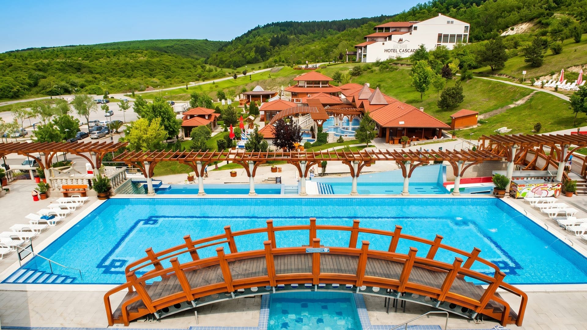 Aerial view of the pools with a wooden bridge, pergolas and the hotel on the hillside