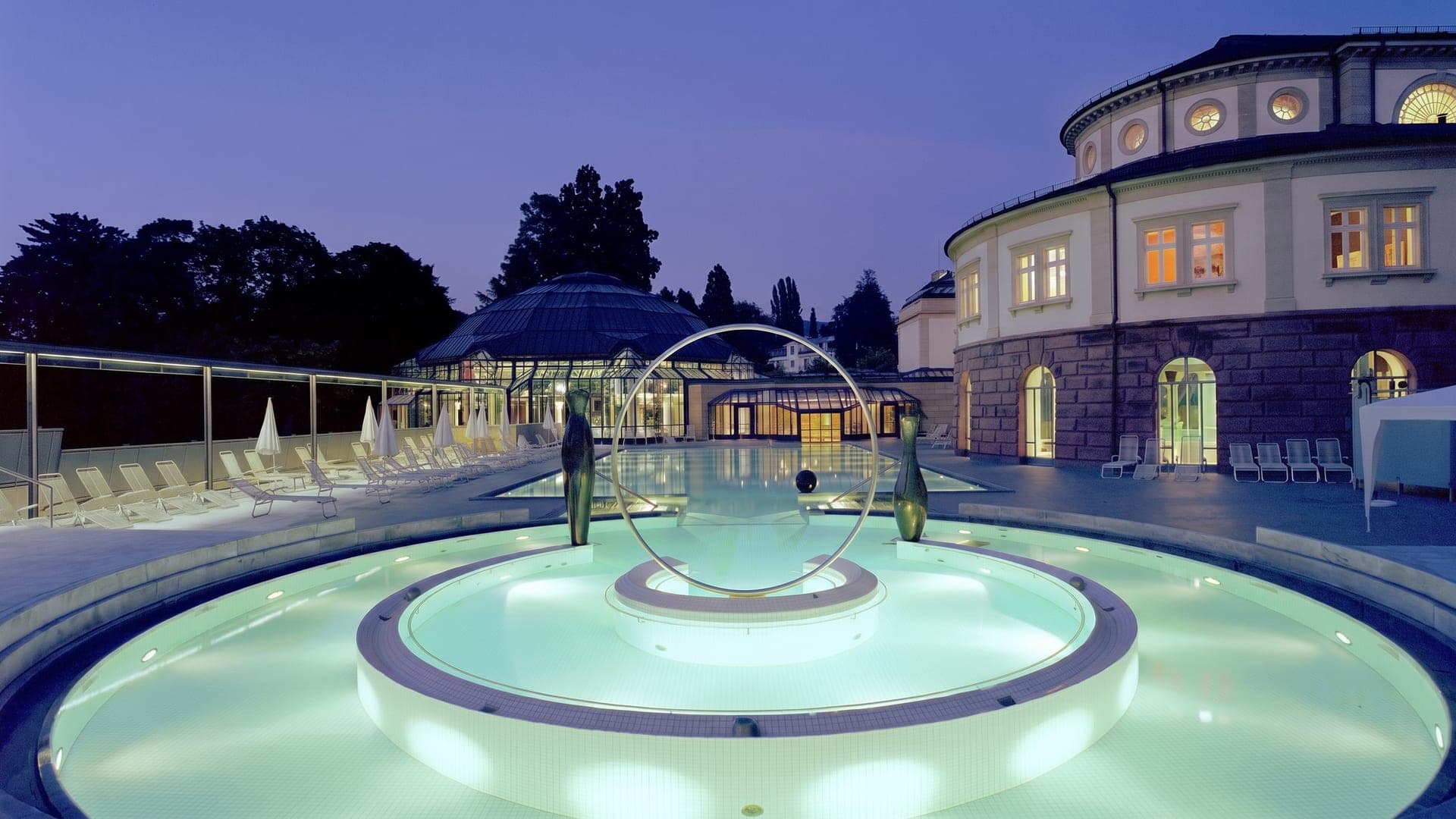 Illuminated circular pool feature at dusk in front of a domed historic bath building
