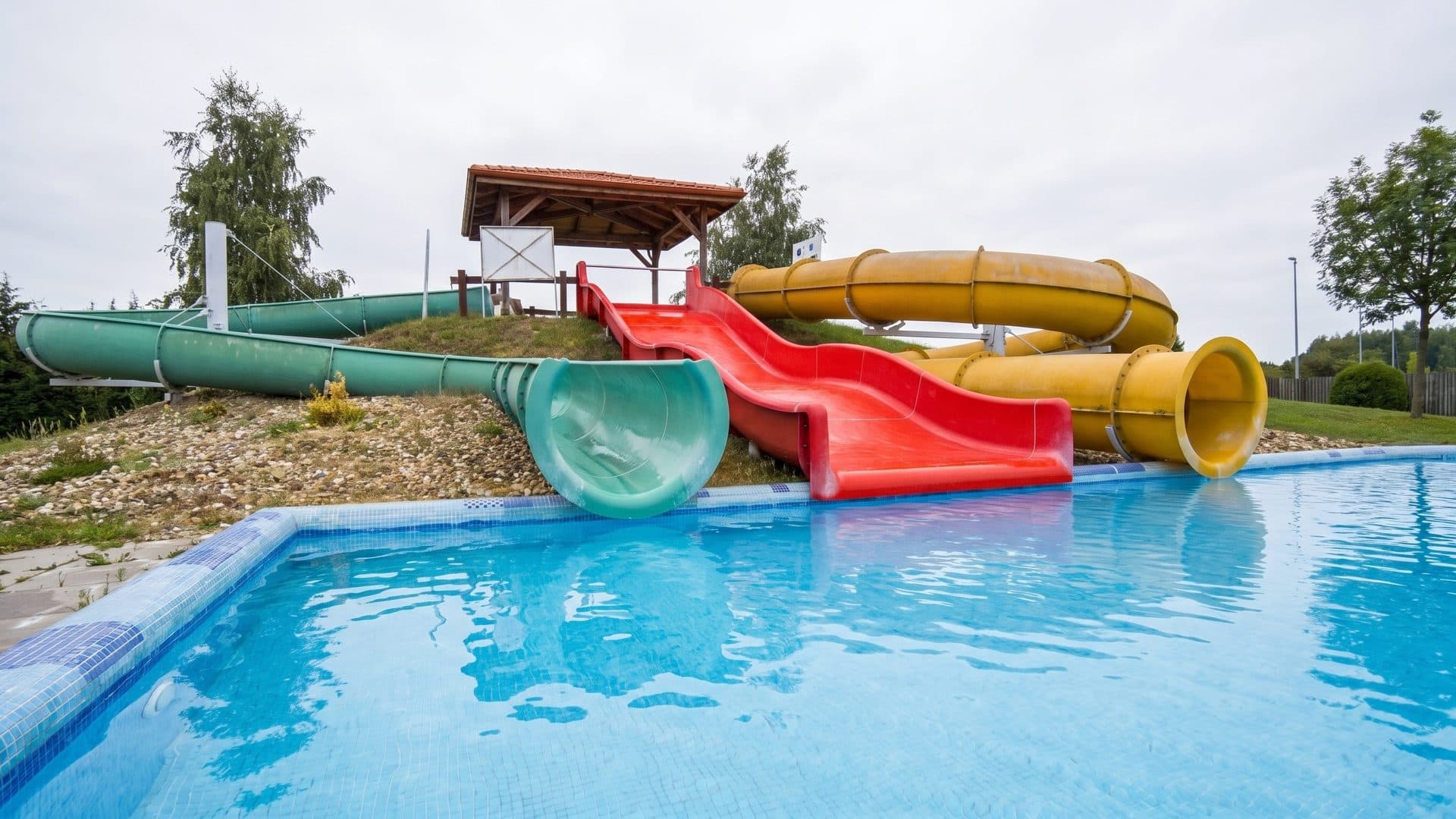 Green, red and yellow waterslides coming down a grassy mound into a blue outdoor pool
