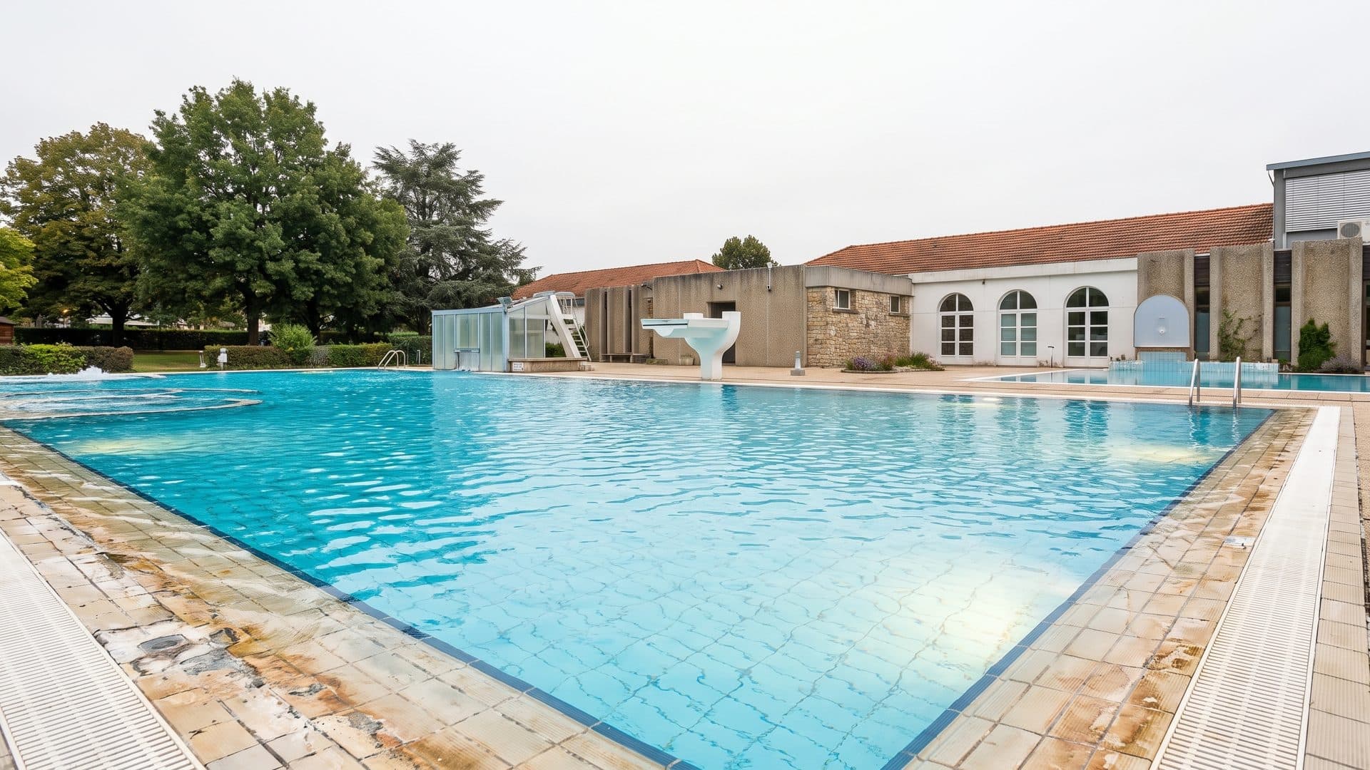 The outdoor thermal pool at Yverdon-les-Bains in full blue water, with the arcaded bath buildings and mature trees along the far side