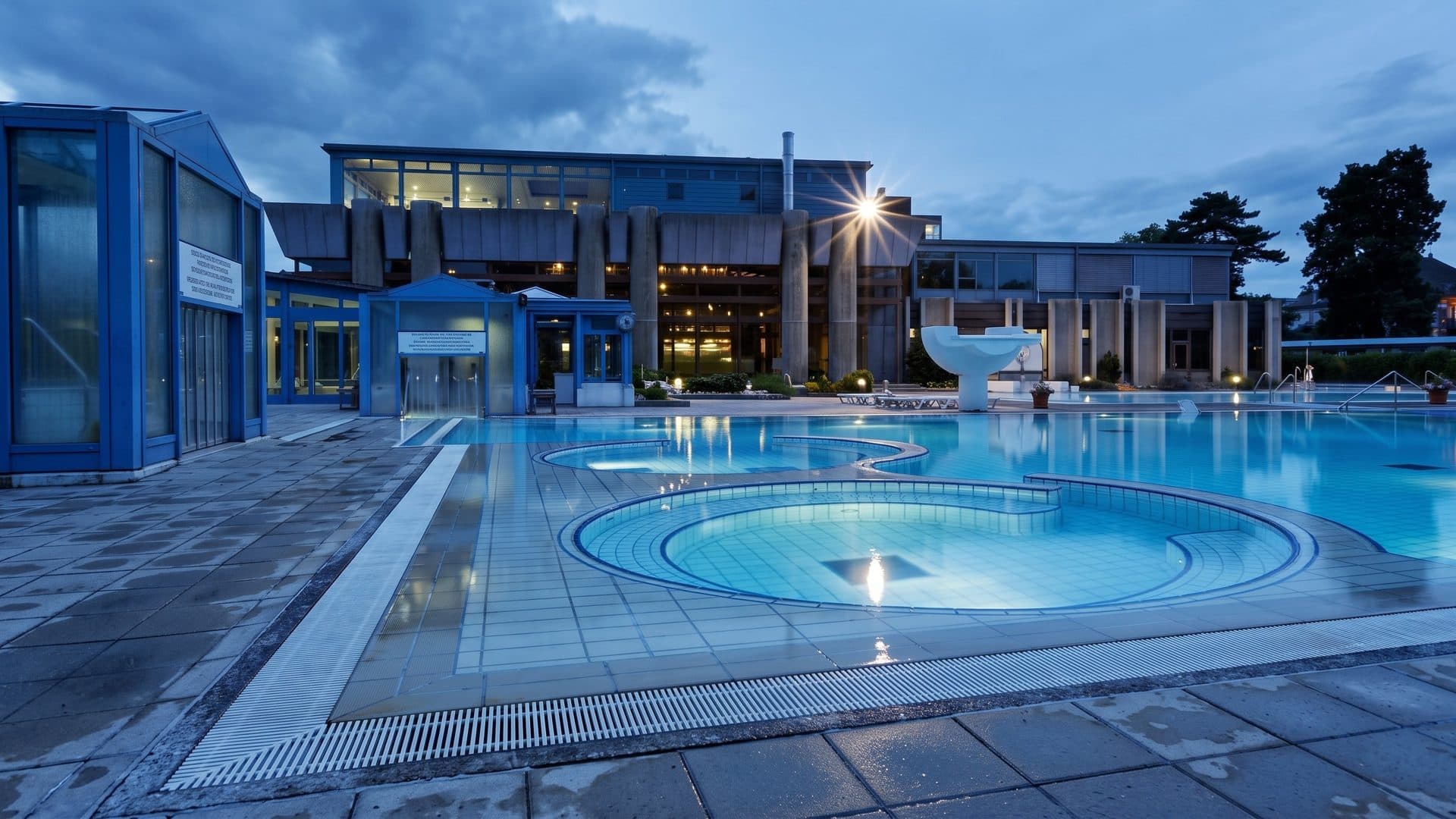 The outdoor thermal pool of Bourbonne-les-Bains with round whirlpool basins, the lit bath building behind at dusk