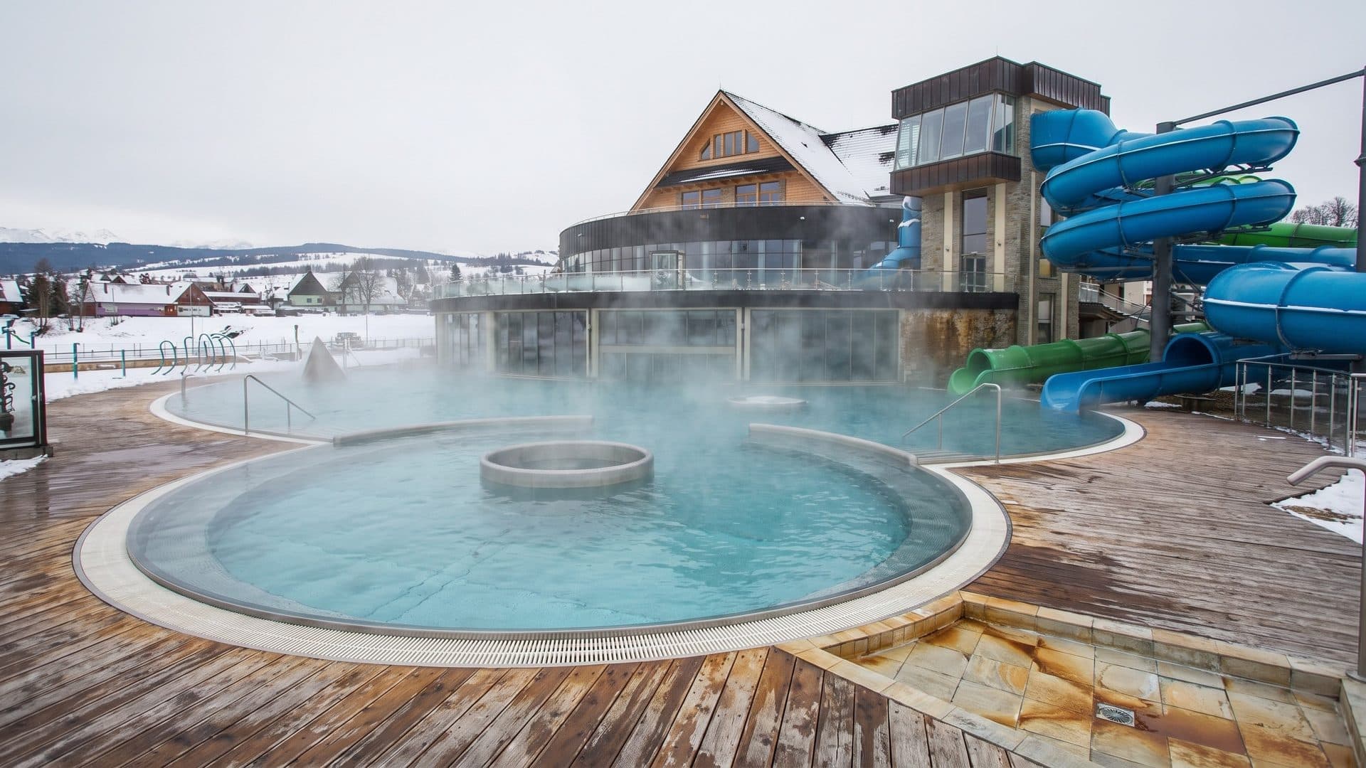 Steaming round outdoor thermal pool on a timber deck, with the mountain-style lodge and blue slides behind in winter