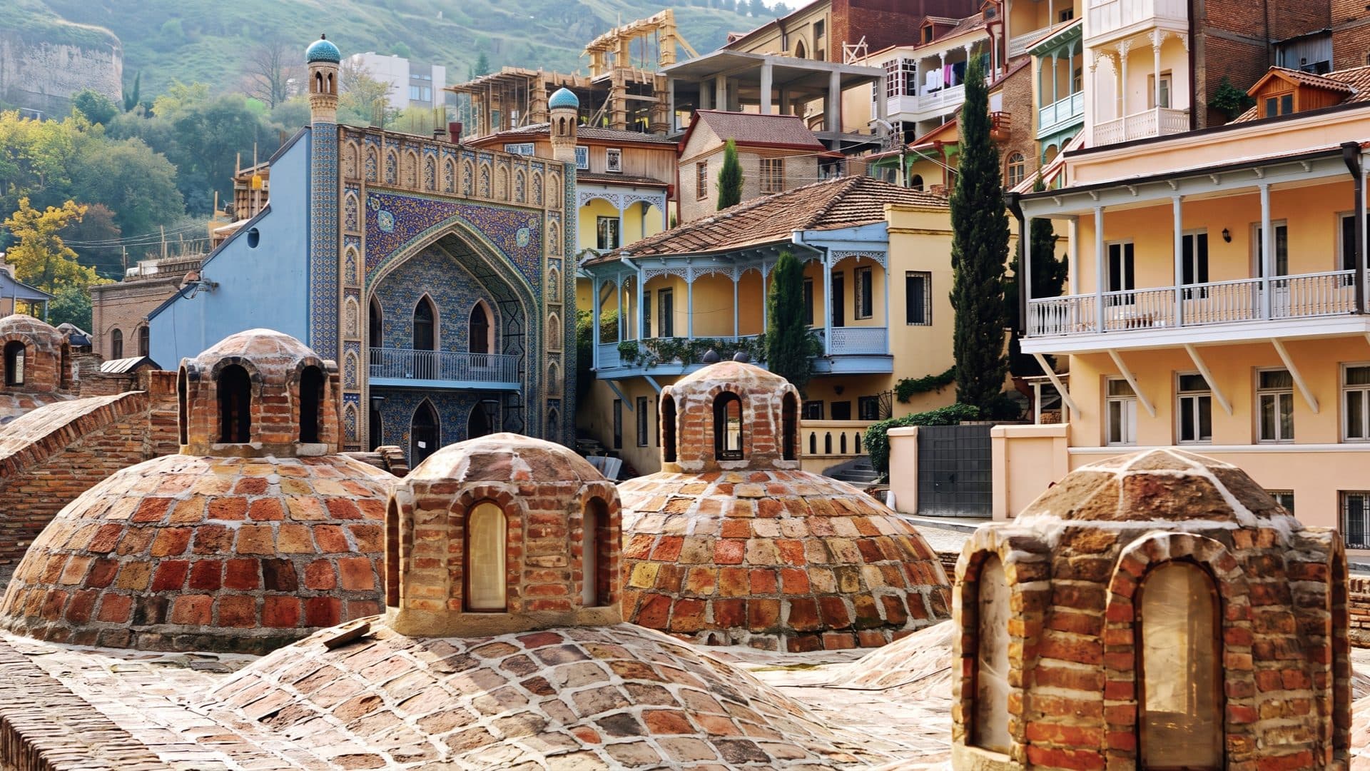 Brick bathhouse domes in Tbilisi’s Abanotubani quarter, with the blue-tiled Orbeliani facade and carved-balcony old-town houses rising behind.