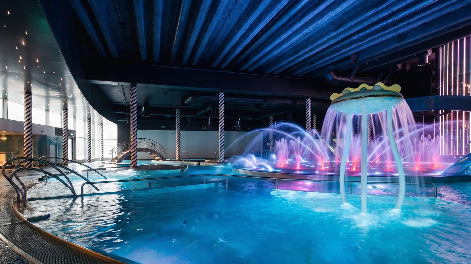 The fountain pool at Cirque de Saimaa under its dark ribbed ceiling, coloured jets arching over the blue water
