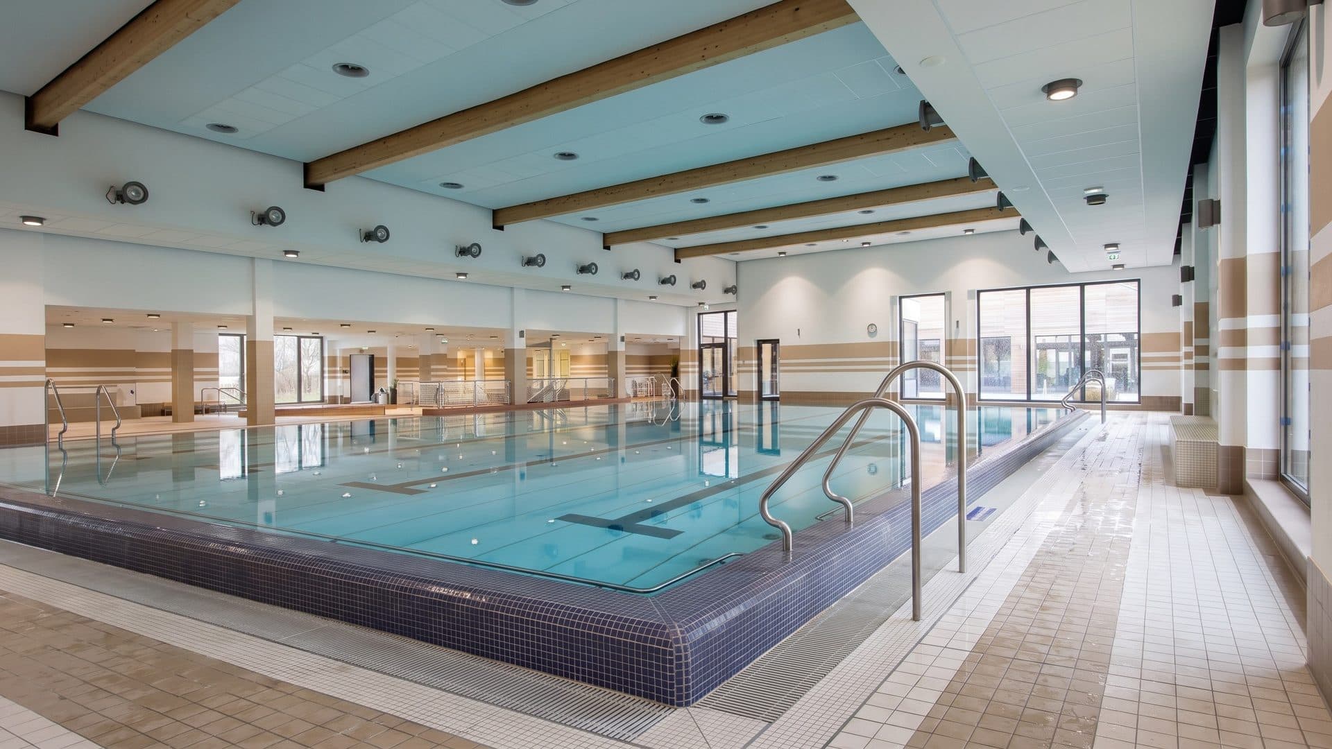 Indoor lap pool with a tiled deck, pale timber ceiling beams and a row of windows