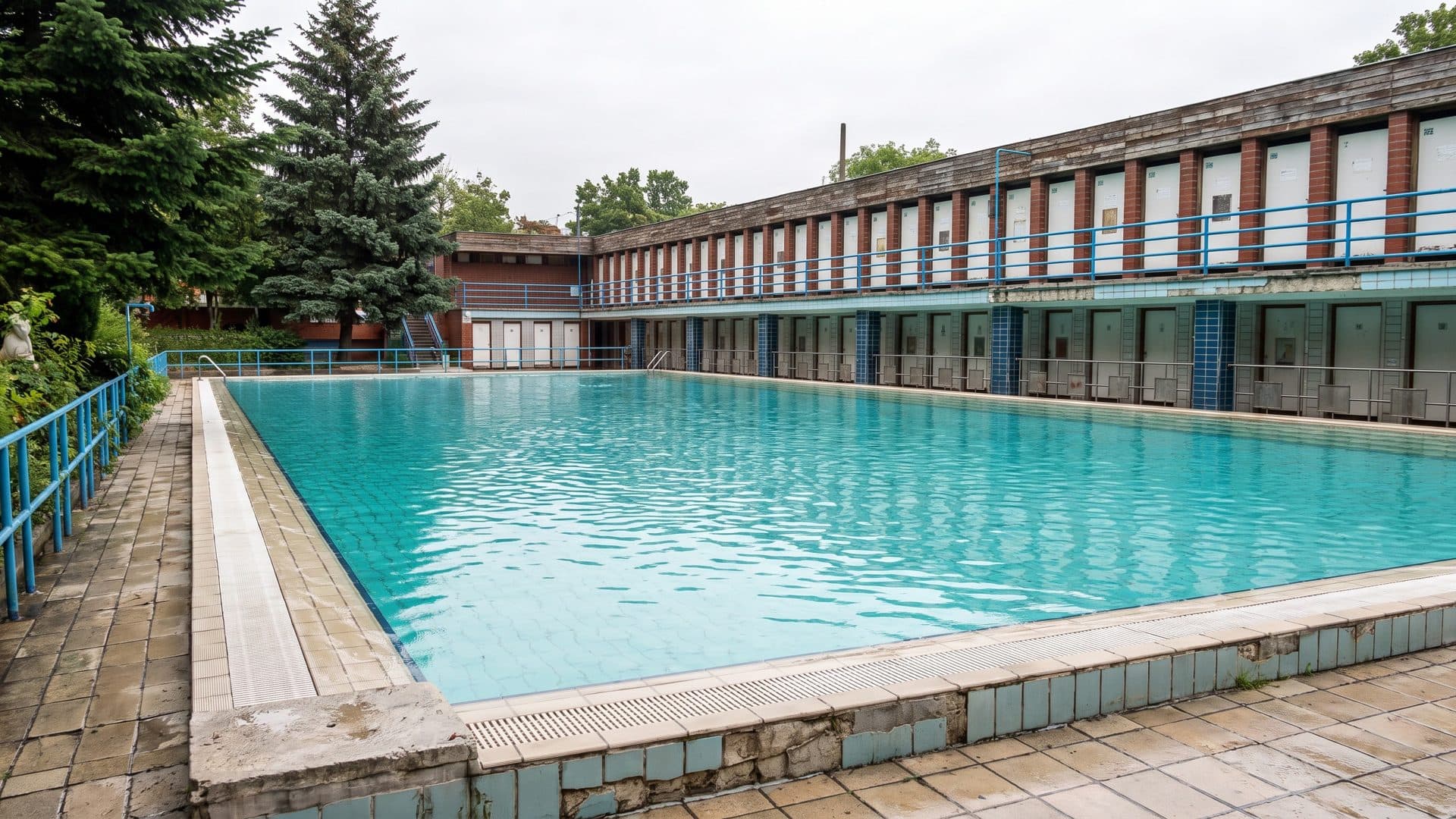 The long outdoor pool in front of the historic two-storey changing-cabin building, framed by spruce trees