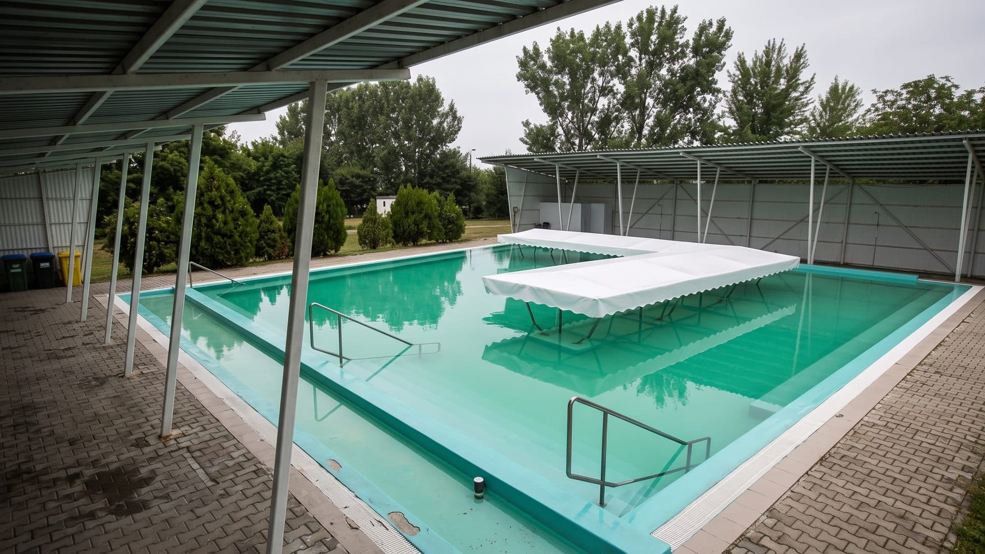 The open-air thermal pool of the Csorna baths with a white shade canopy over the water and a covered stand behind