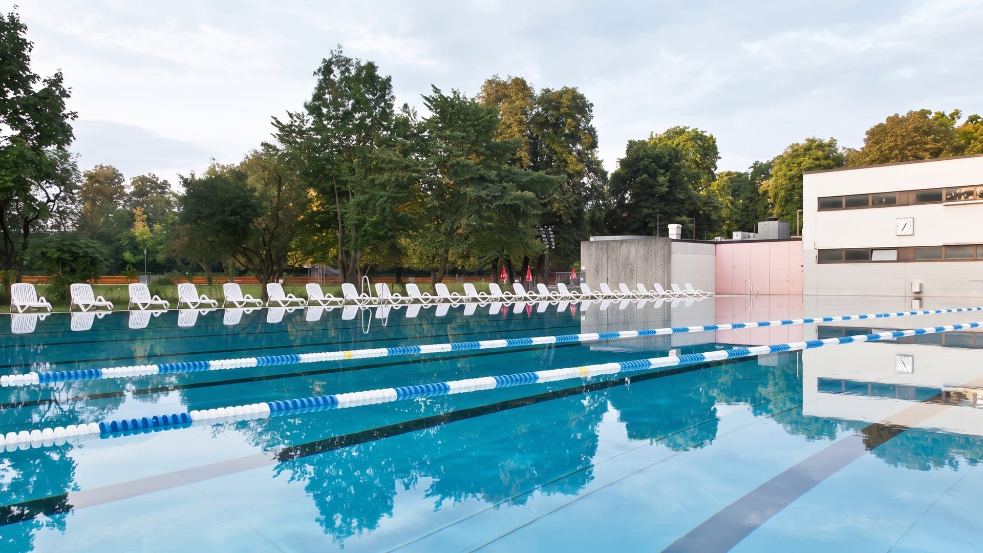 Outdoor lane pool with rows of white loungers beside a modernist building