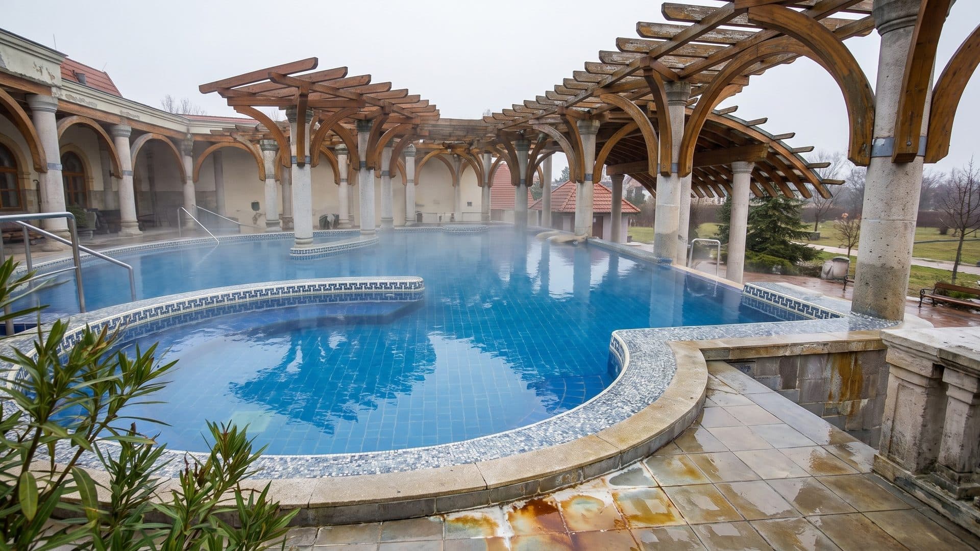 Steaming thermal pool beneath curved timber pergolas and an arcaded colonnade
