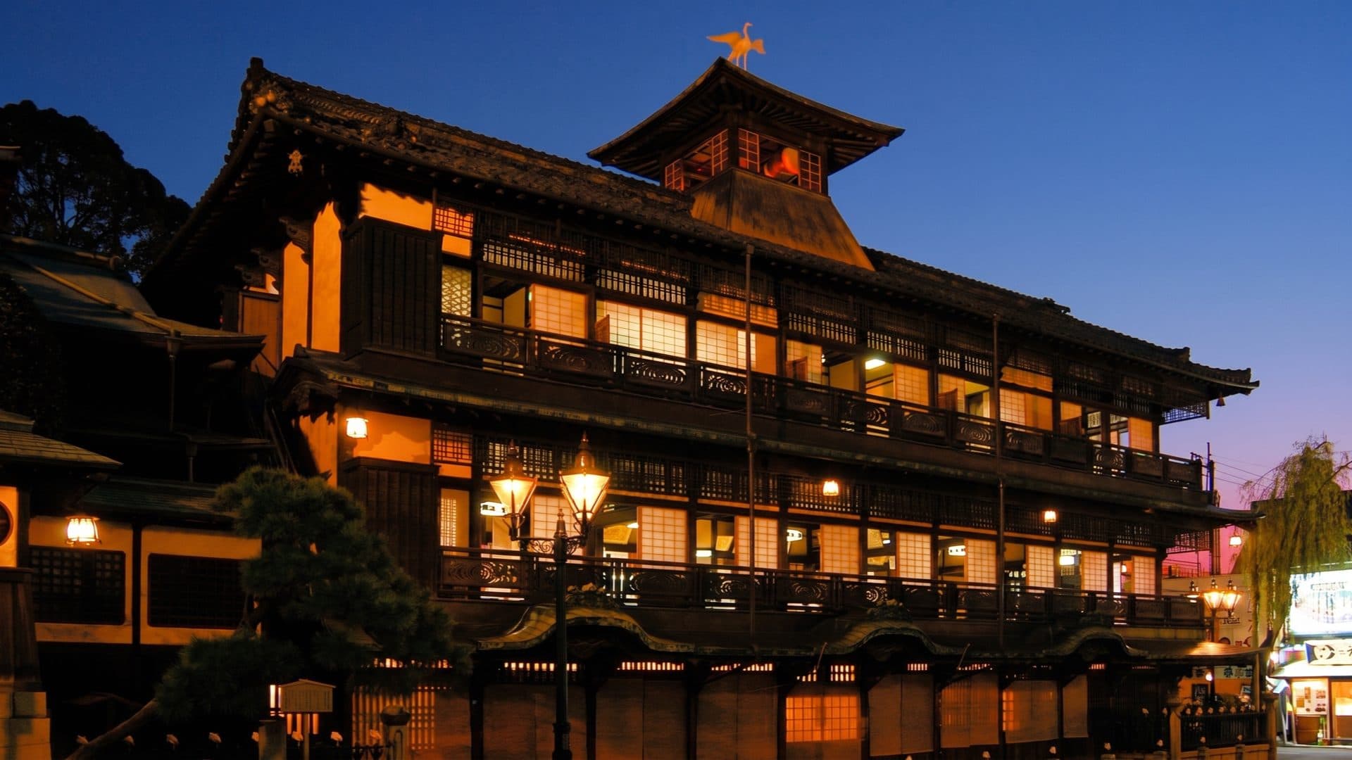 The historic three-storey wooden Dogo Onsen bathhouse lit at dusk, its heron-topped turret rising against a deep-blue sky.
