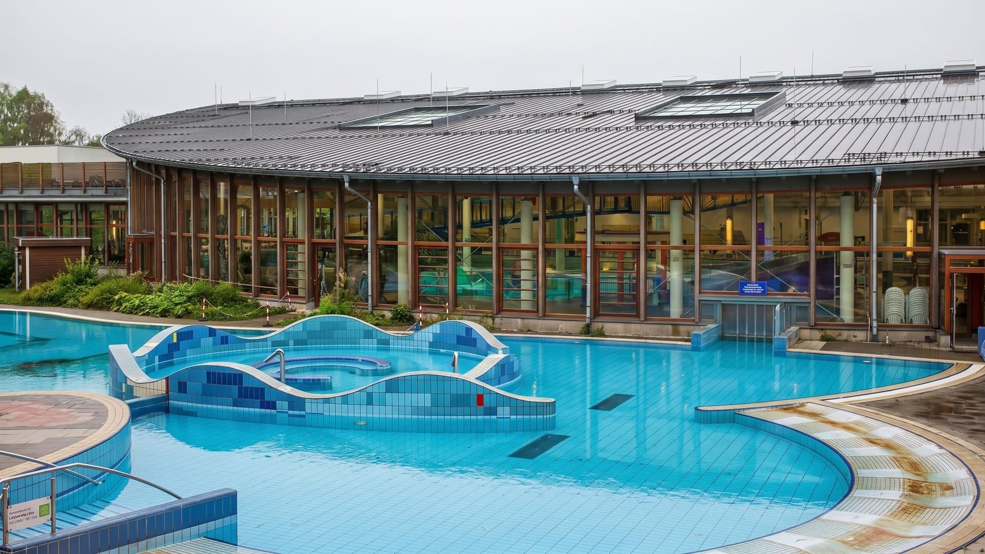 Outdoor leisure pool with a mosaic-tiled wave bench, in front of the glazed pool hall with its long curved roof
