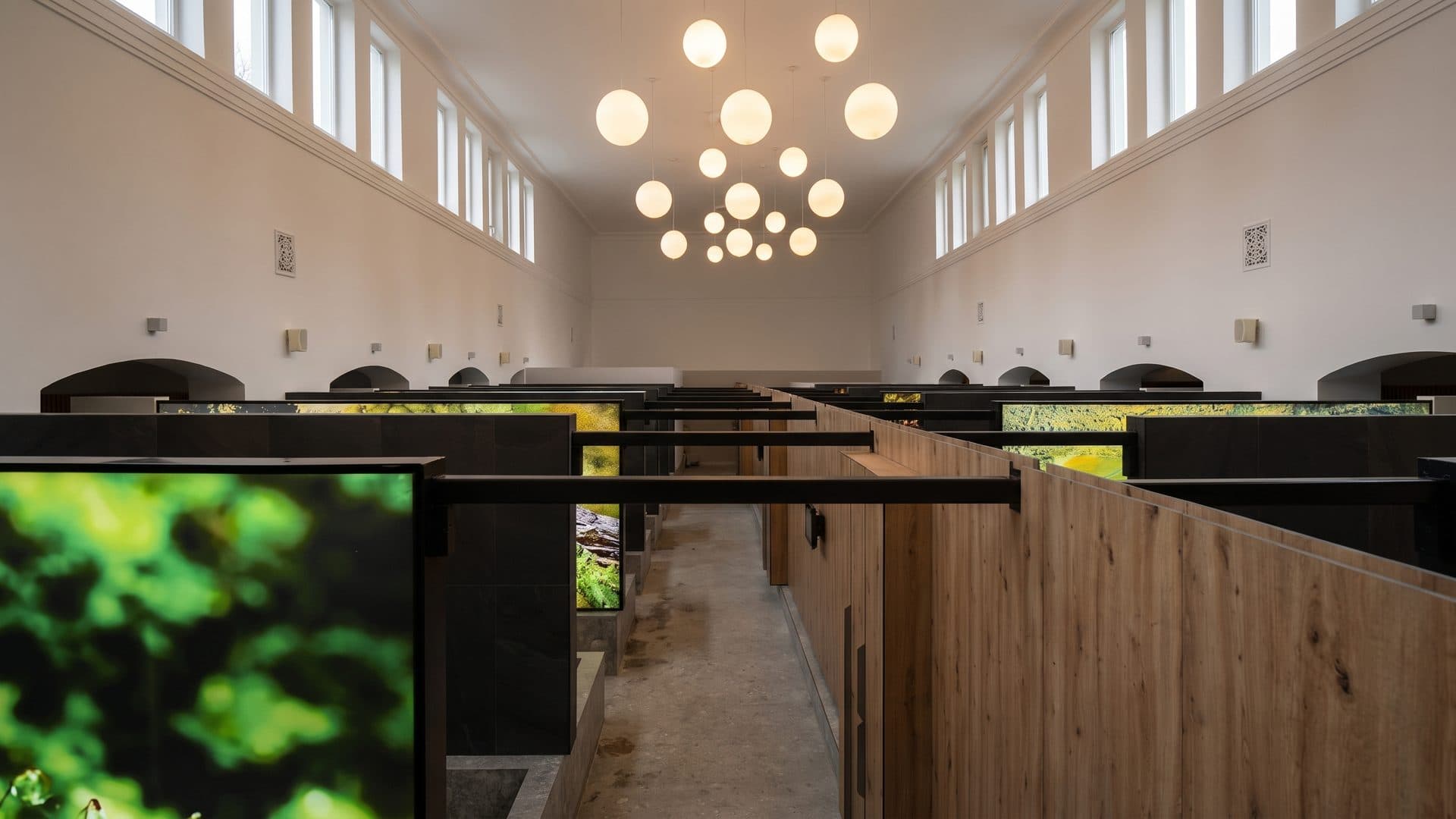 The treatment hall at the Druskininkai sanatorium, rows of dark tiled bath cabins under a high white ceiling with cluster pendant lights and lit greenery panels along the wall