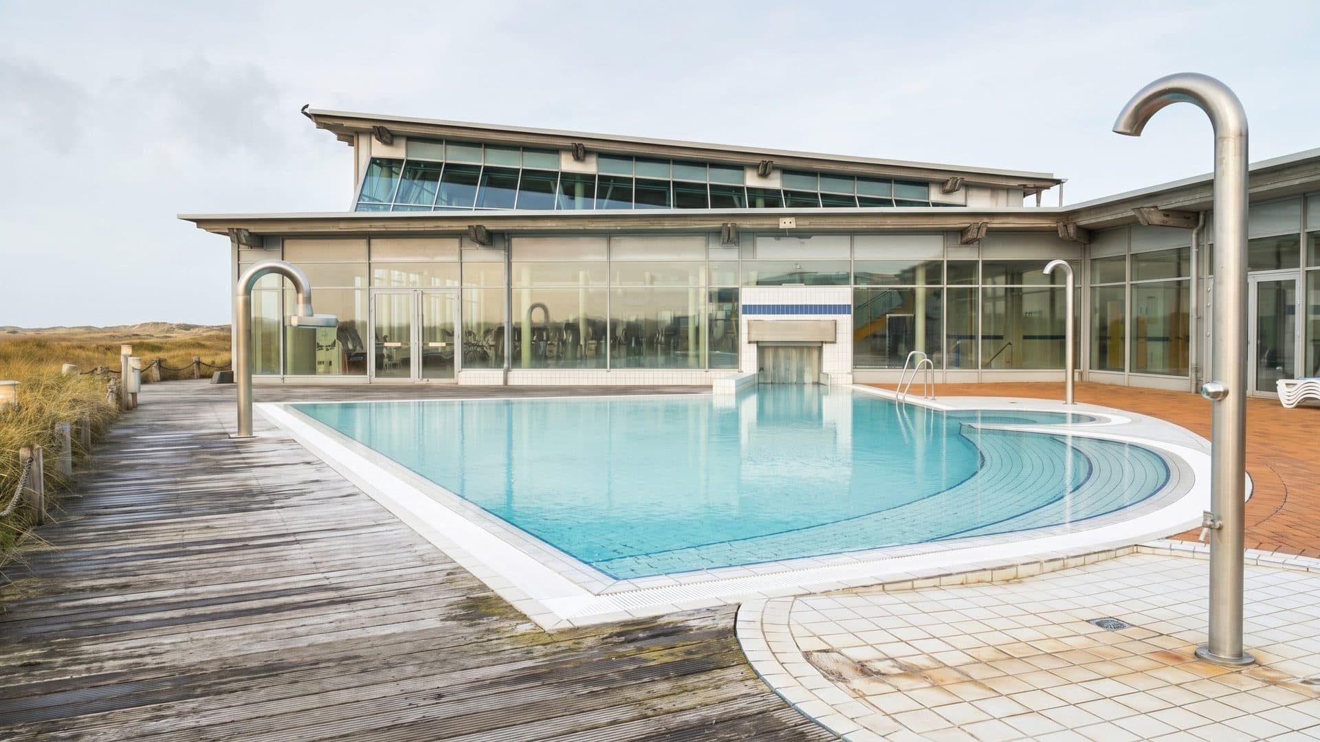 Outdoor thermal pool on a timber deck among the dunes, with steel shower posts and the glazed bath house