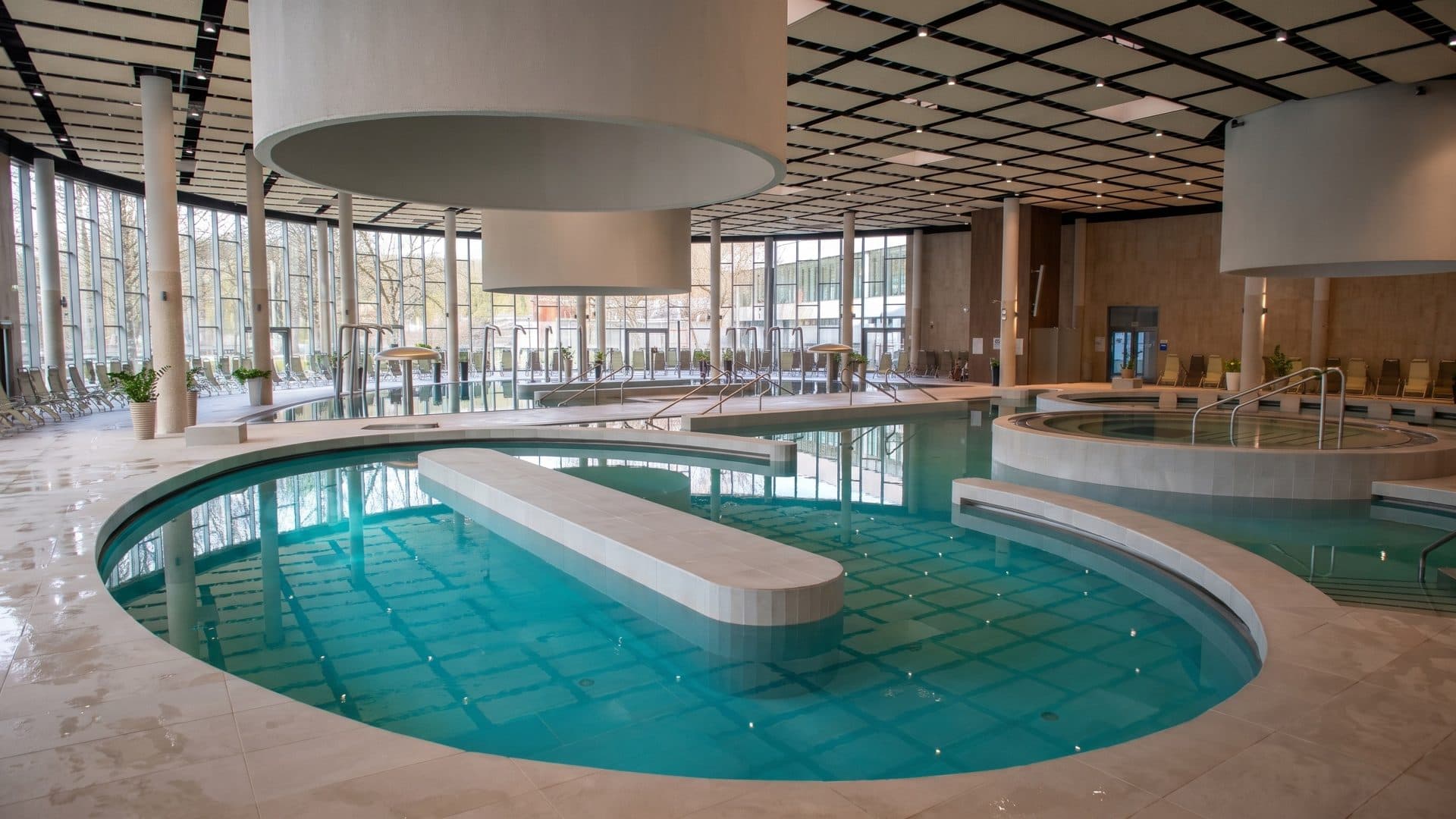 Oval indoor thermal pool with a central lounging bench, under large round ceiling lamps in a wide glazed hall