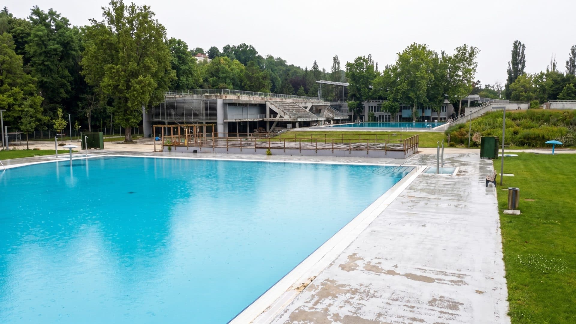 Large open-air pool with a wide concrete deck, lawns and the terraced stands of the complex beyond