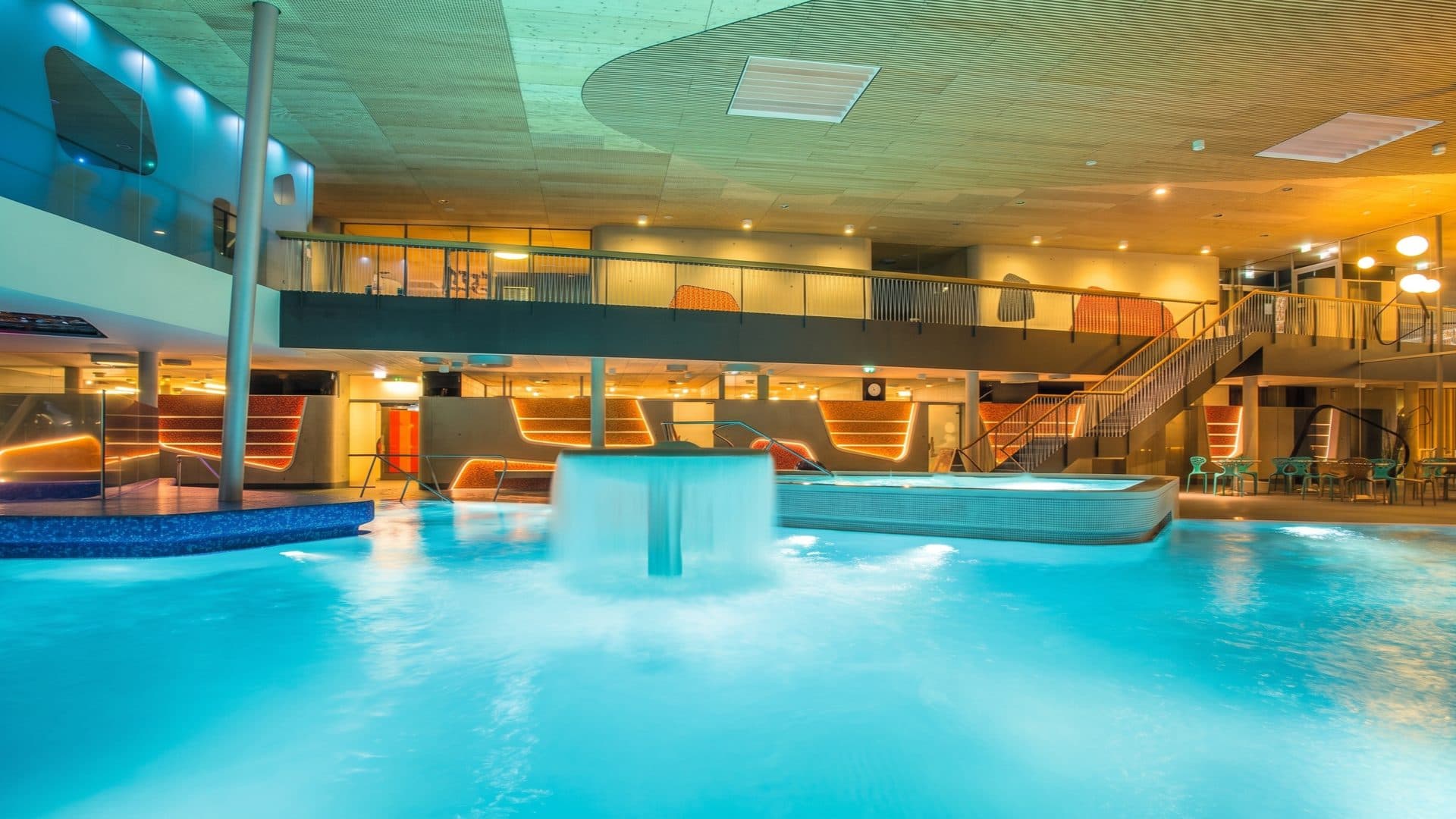 Modern indoor thermal pool hall with a mushroom fountain spilling into turquoise water, a warm timber ceiling and orange-lit lounger niches on two levels.