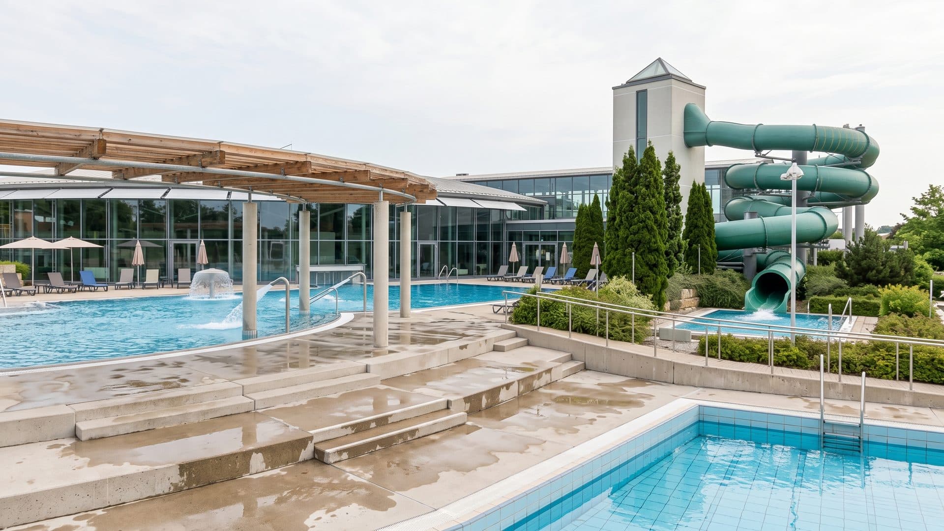 Outdoor pools in front of the glass hall, with the green tube slides rising beside the building.