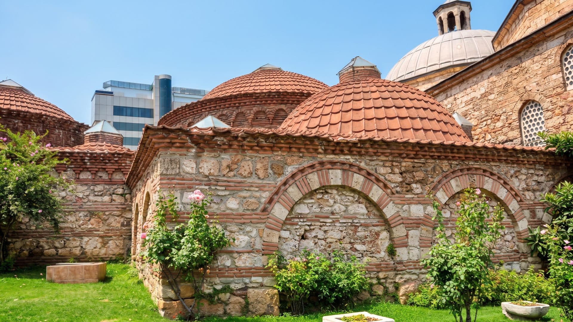 The domed stone-and-brick exterior of a historic Ottoman bathhouse, its tiled cupolas rising over a rose garden.