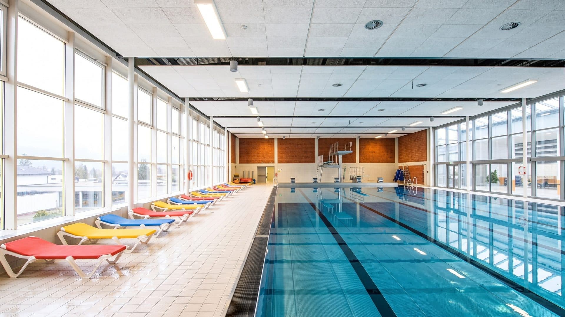 The lane pool of the espada Freizeitbad along a wall of tall windows, coloured loungers on the tiled deck and a diving board at the far end
