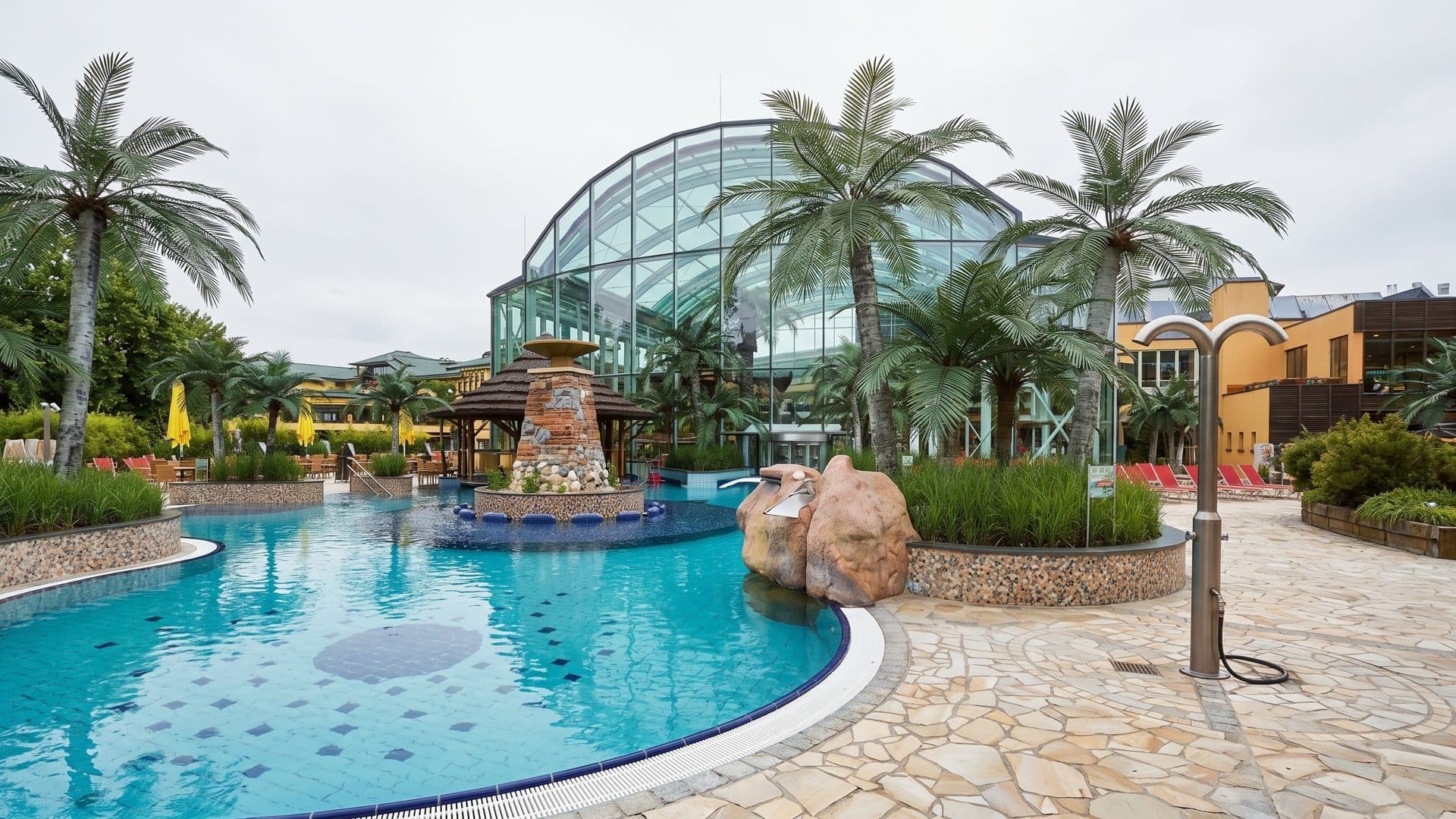 Outdoor thermal pool among palm trees, with a stone fountain island and the glass dome of the palm house