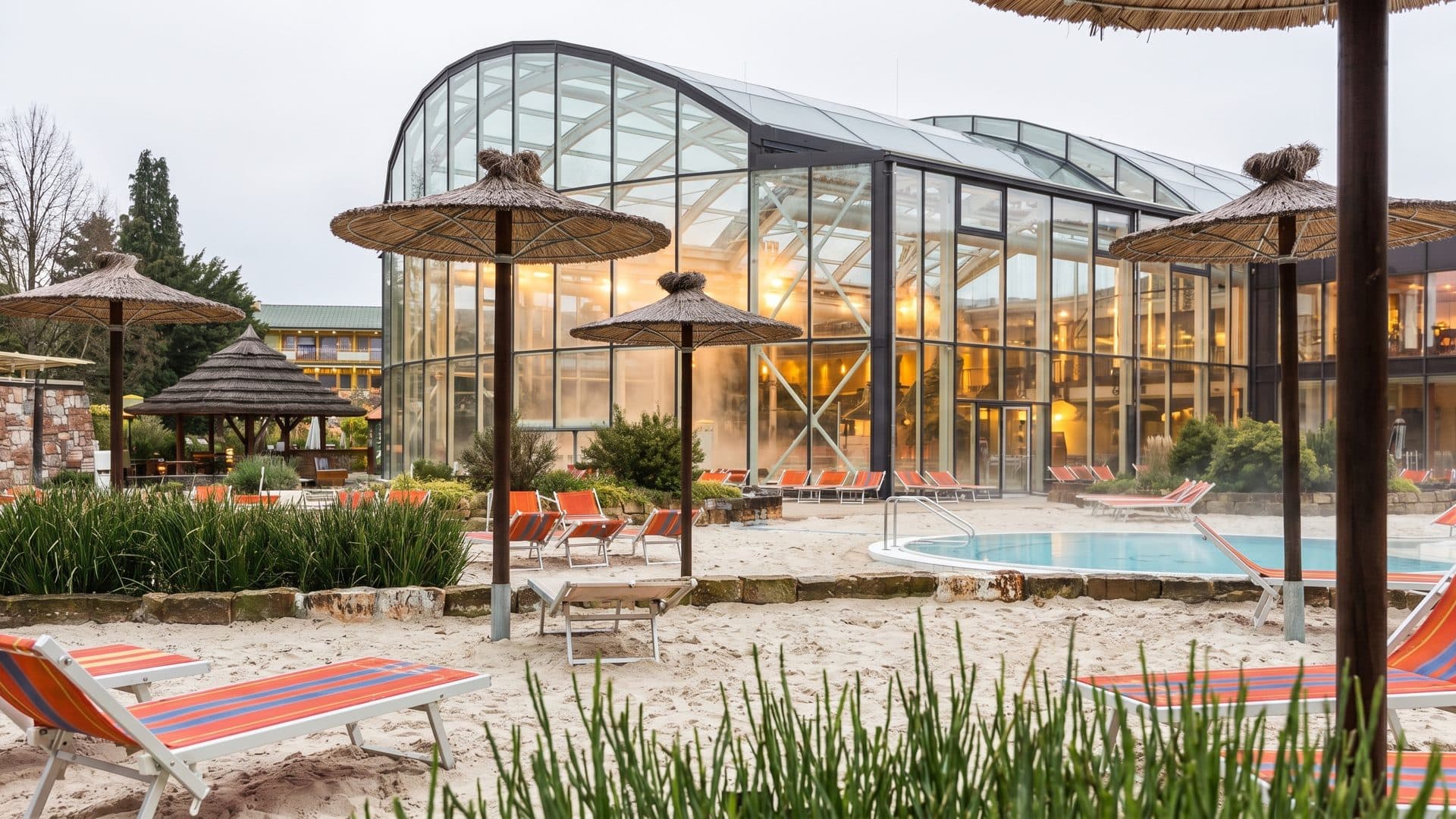 The glazed hall of the Eurotherme in Bad Schallerbach seen across the outdoor pool, with parasols and loungers on the deck