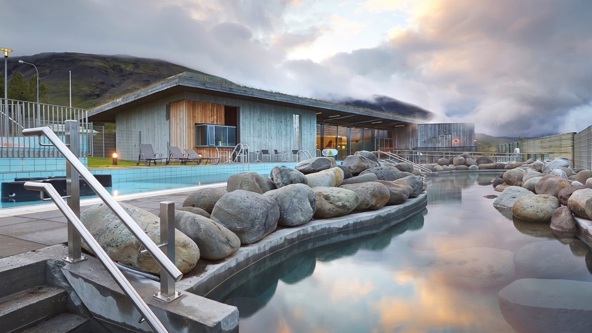 Turquoise pool and a steaming lakeside hot pool bordered by rounded boulders, beside a turf-roofed spa building with mountains behind.