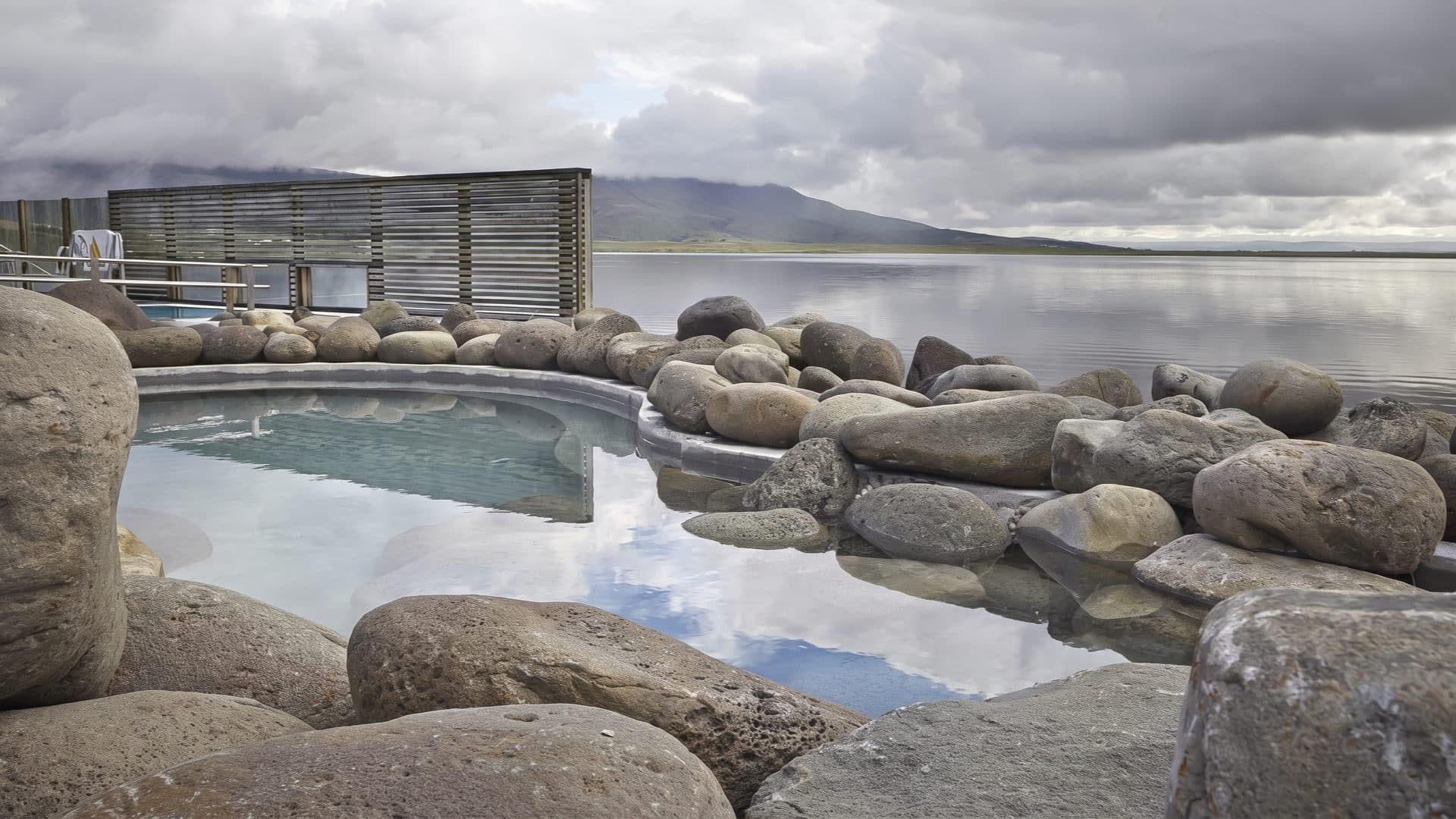 Geothermal bathing pool ringed with boulders at the edge of a calm lake, a timber screen and low mountains beyond under a grey Icelandic sky.