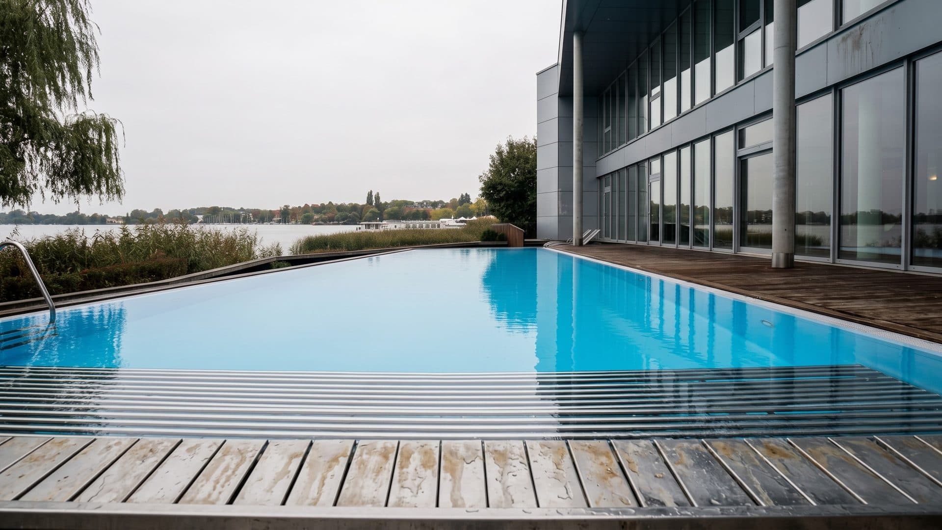 The infinity-edge outdoor pool of the Fontane-Therme on the shore of Lake Ruppin, a timber deck in front and the glass building beside it