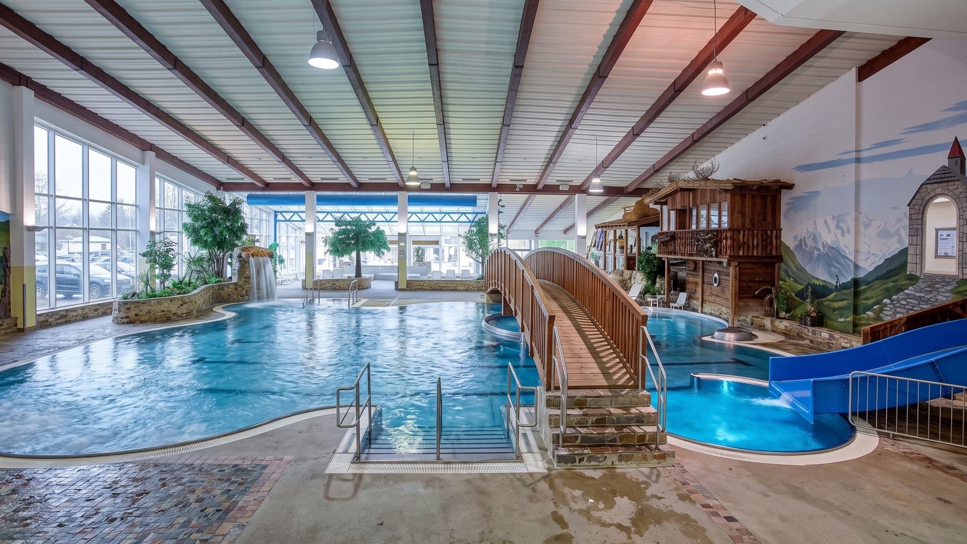 The indoor leisure pool of the Freizeit Arena Solden with a timber bridge, a blue slide and an alpine mural