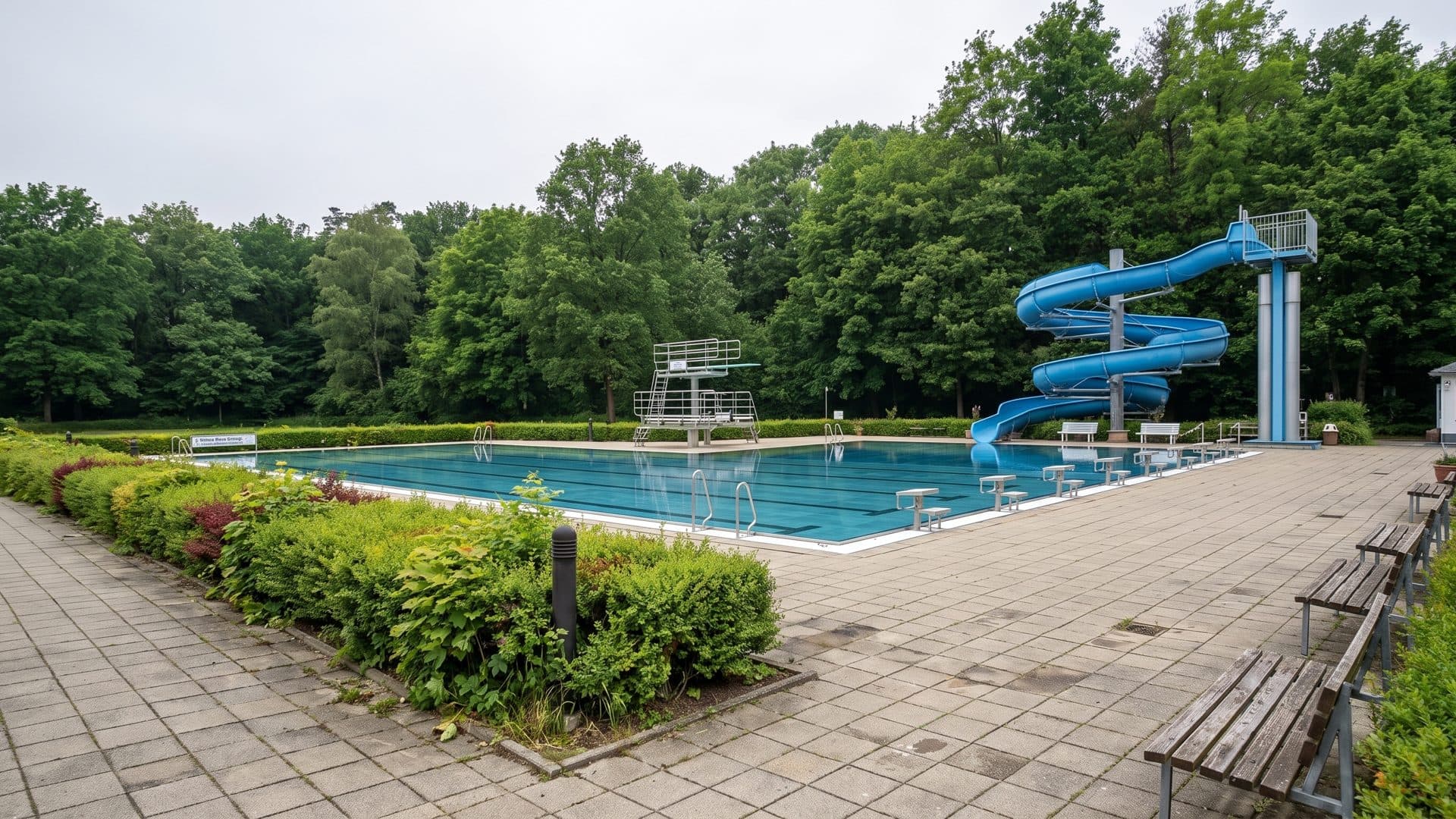 The open-air pool at Freizeitbad Billy with a blue water slide curling down at one end, clipped hedges around the deck and forest behind