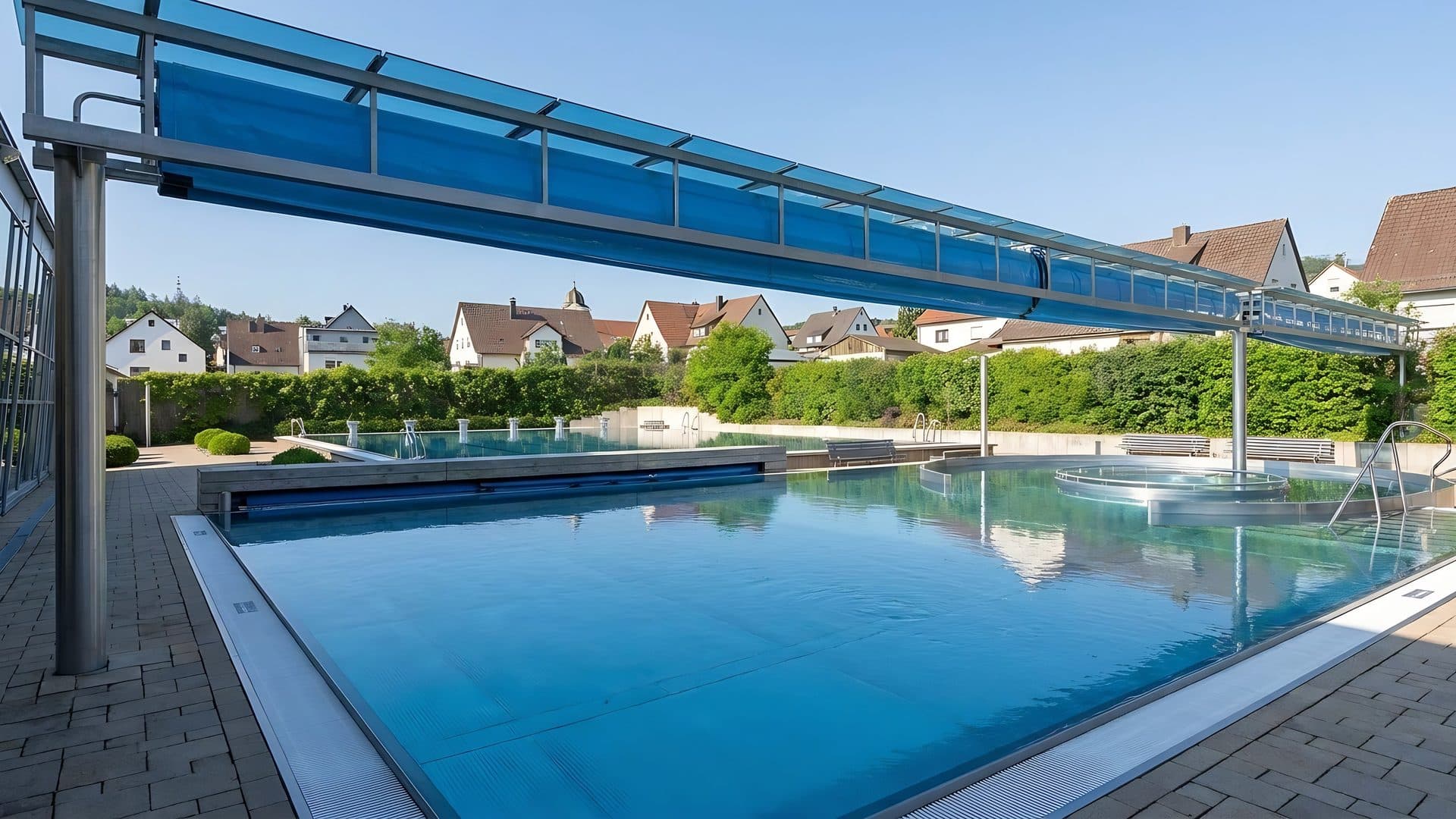 The outdoor pool of Freizeitbad Nautilust under its blue glazed canopy with village roofs behind