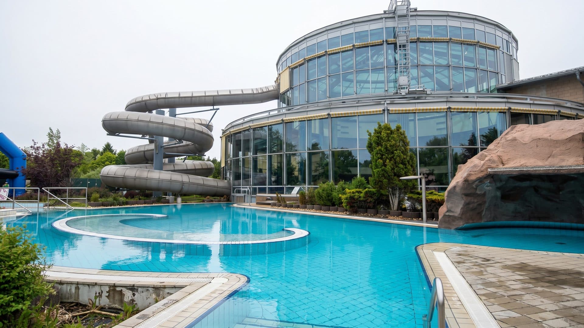 Outdoor leisure pool with a grey spiral slide beside the round glazed hall and an artificial rock