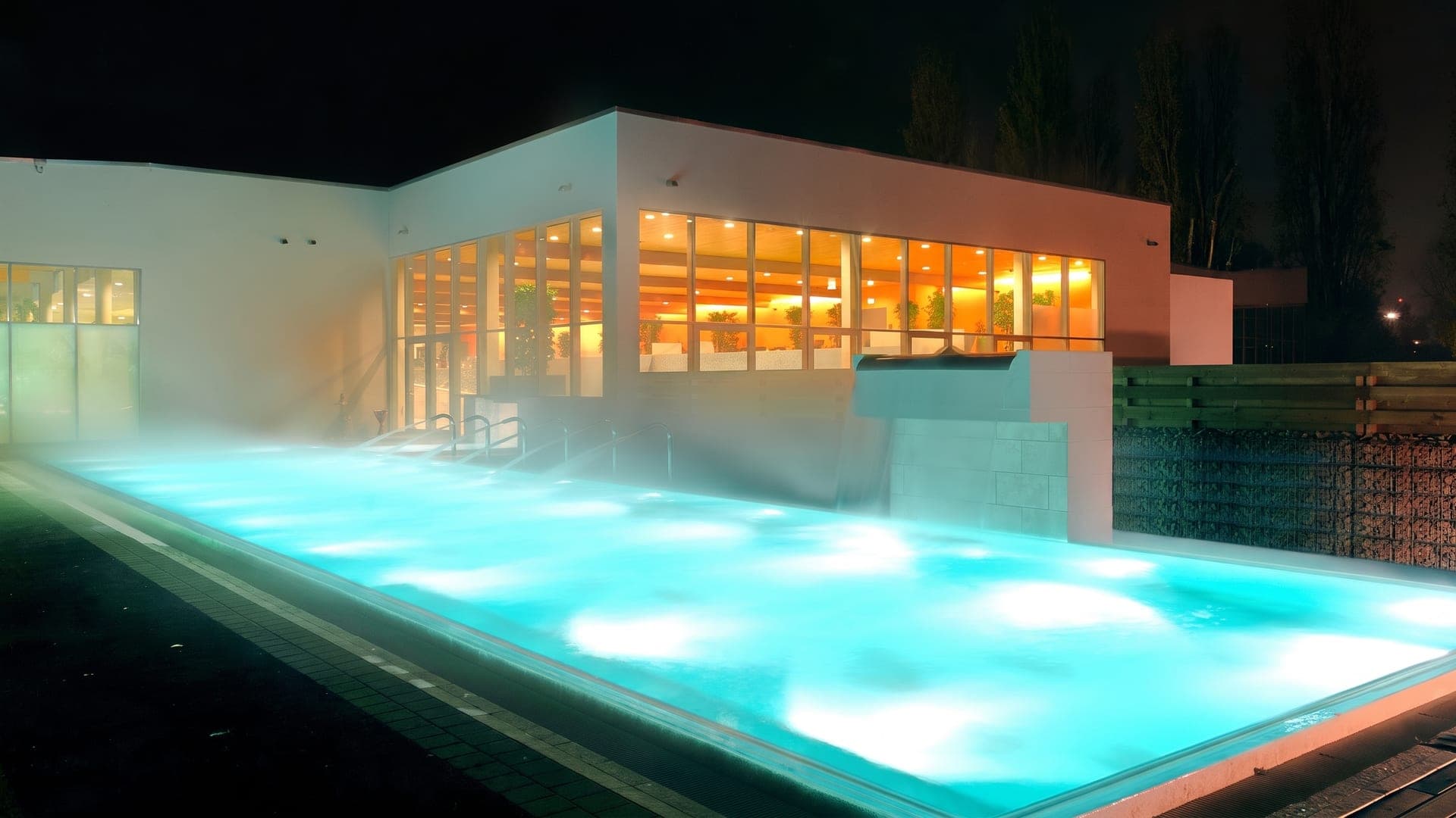 The outdoor pool at Fuerthermare in Fuerth lit at night, steam rising in front of the glowing glass building.