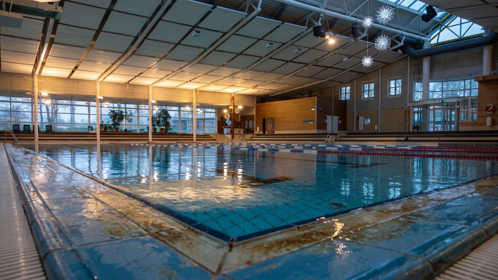 Indoor swimming hall with lane ropes, timber ceiling and a wall of windows down one side.
