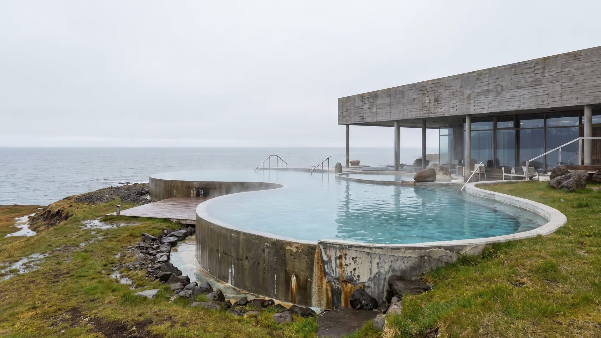 Infinity-edge geothermal pools on the shore, with a concrete bath house and the sea beyond