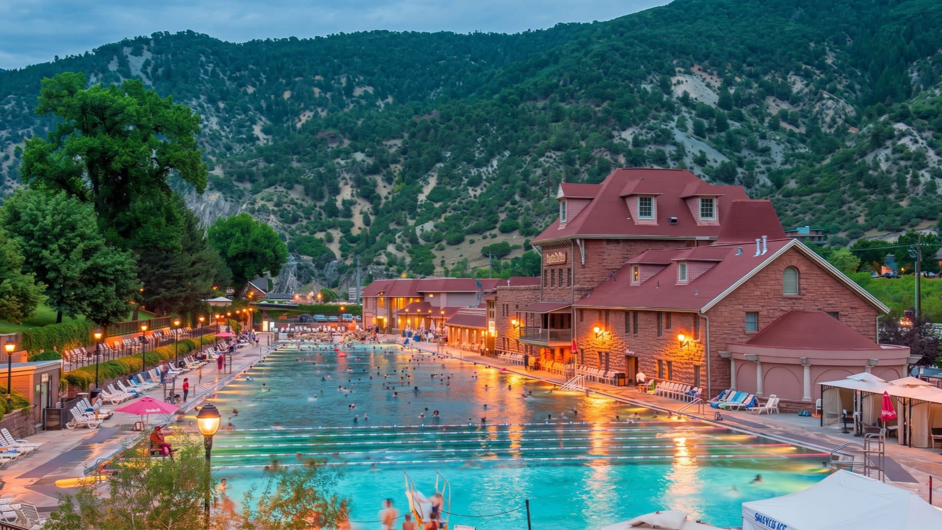 The large historic hot-springs pool at dusk, bathers in the long green-lit water beside a red-roofed sandstone bathhouse, forested canyon walls rising behind.