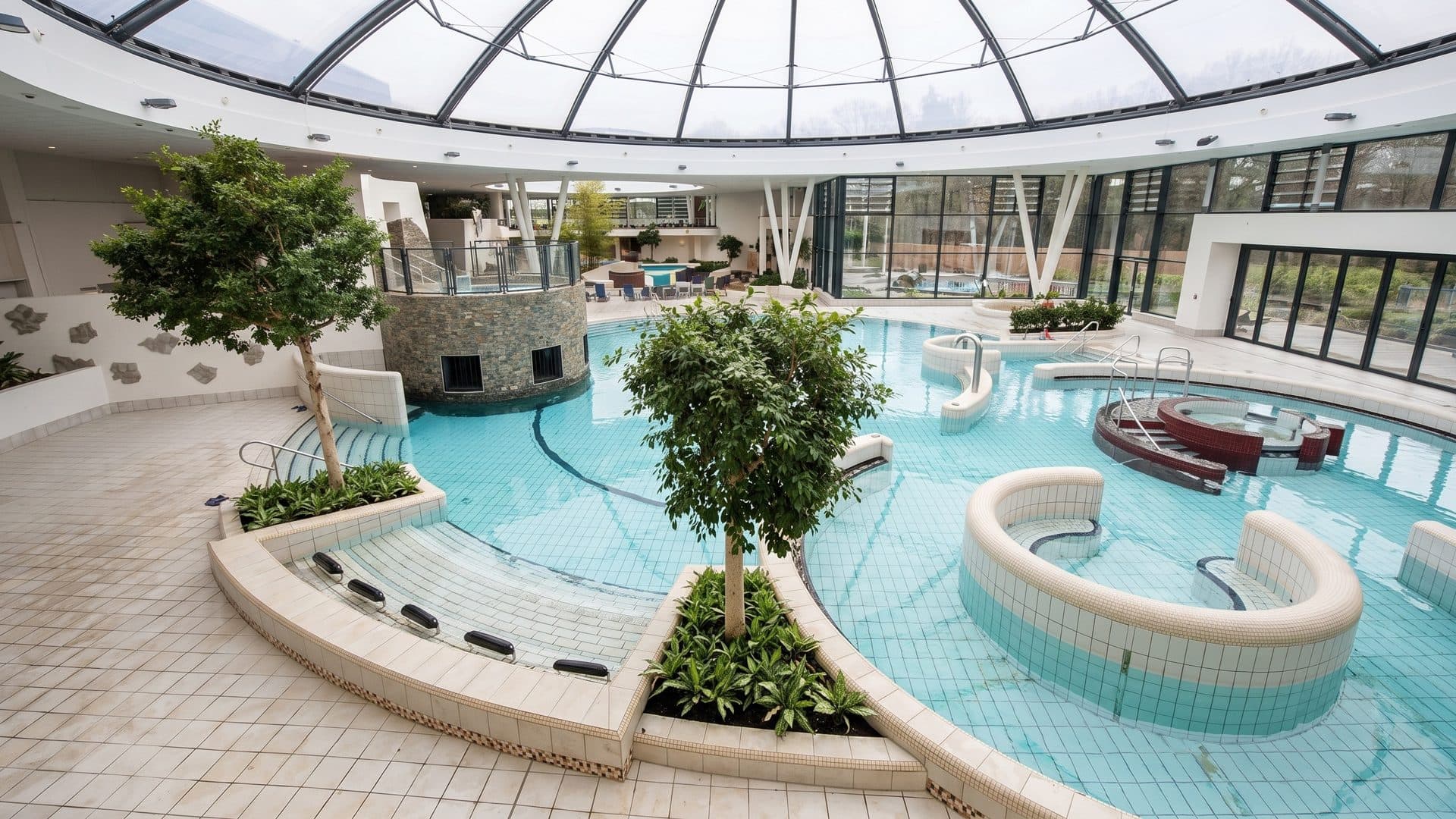 Indoor thermal pool landscape under a glass dome, with round lounging islands and trees in planters
