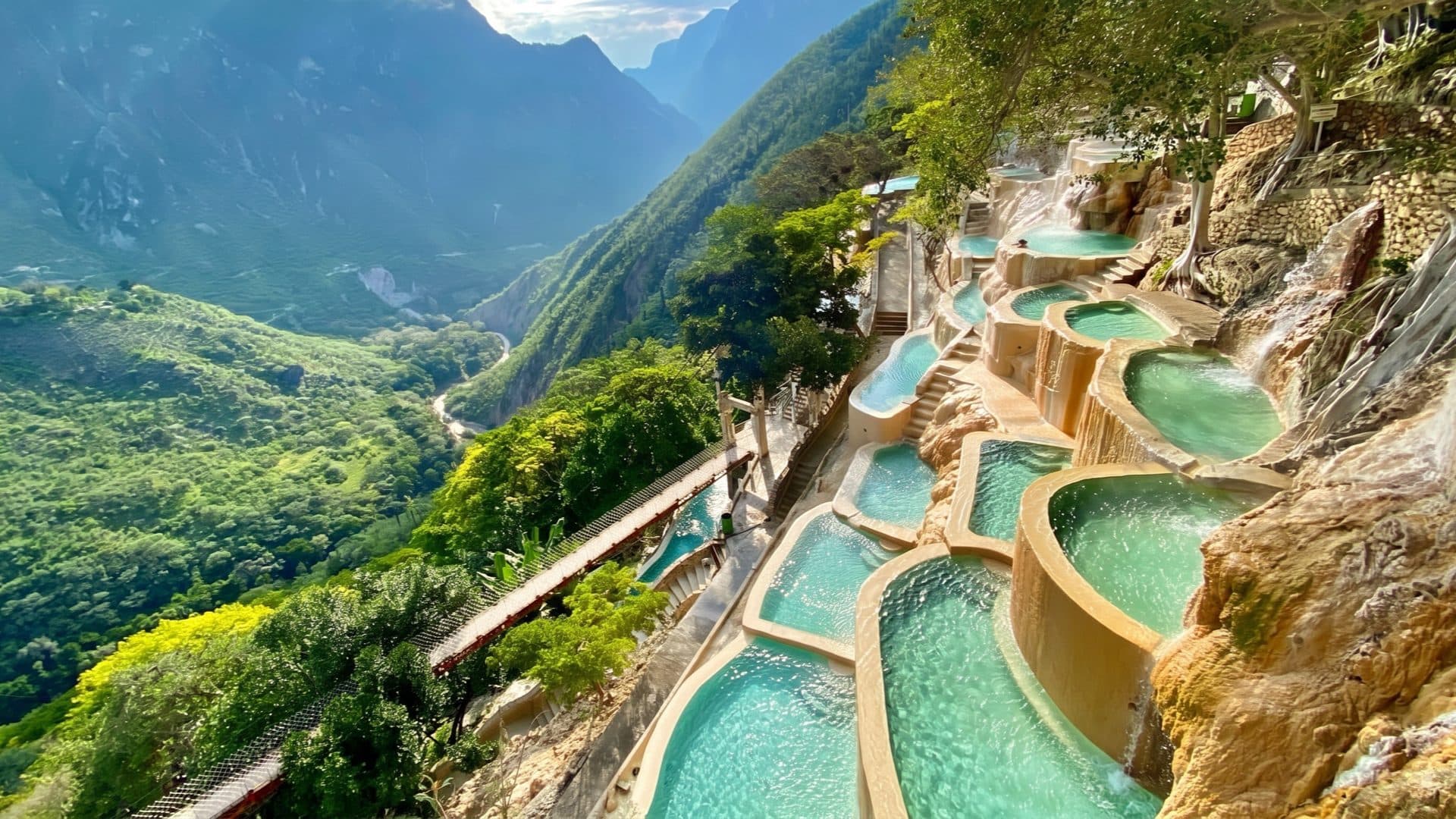 Terraced turquoise pools stacked down a canyon cliff, with a walkway and lush green mountains dropping to a river far below.
