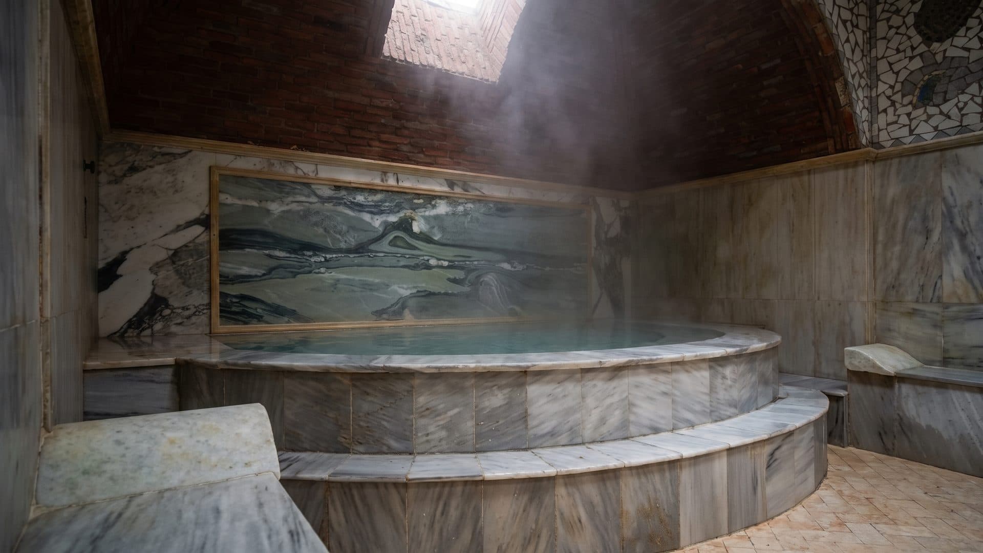 Round marble pool under a brick vault, a long carved marble panel on the wall behind it and steam over the water.