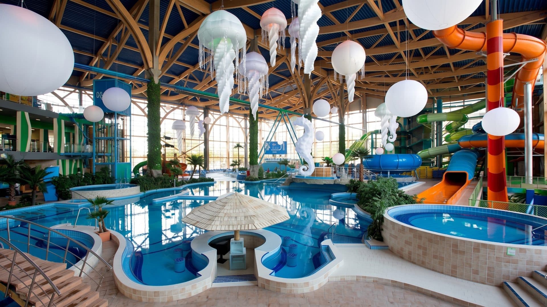 The indoor pools of H2O Park in Rostov-on-Don under a timber-ribbed glass roof hung with jellyfish lamps, slides on the right