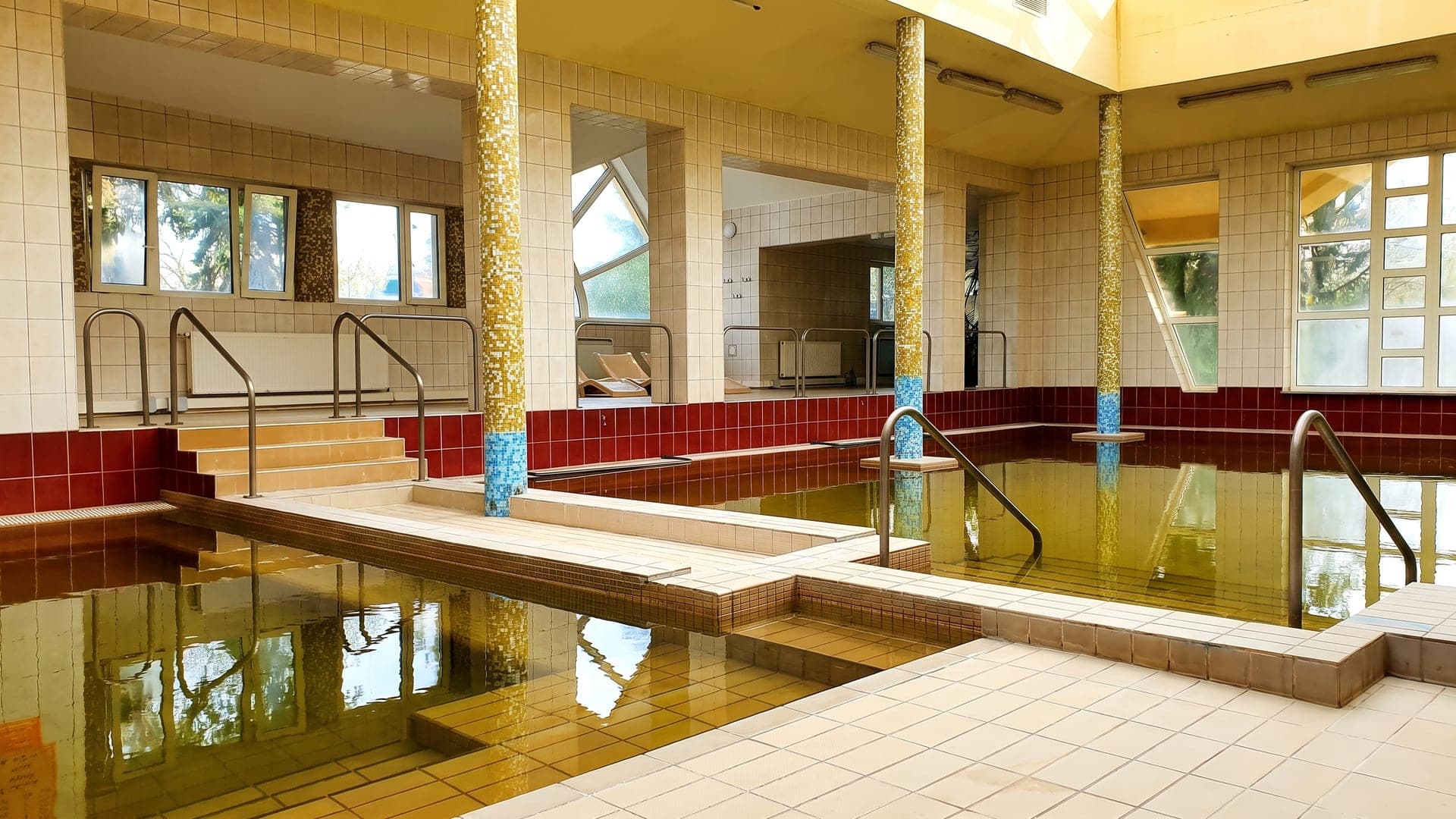 The indoor pools at the thermal bath in Hajdunanas, their dark ochre water between gold-mosaic columns.