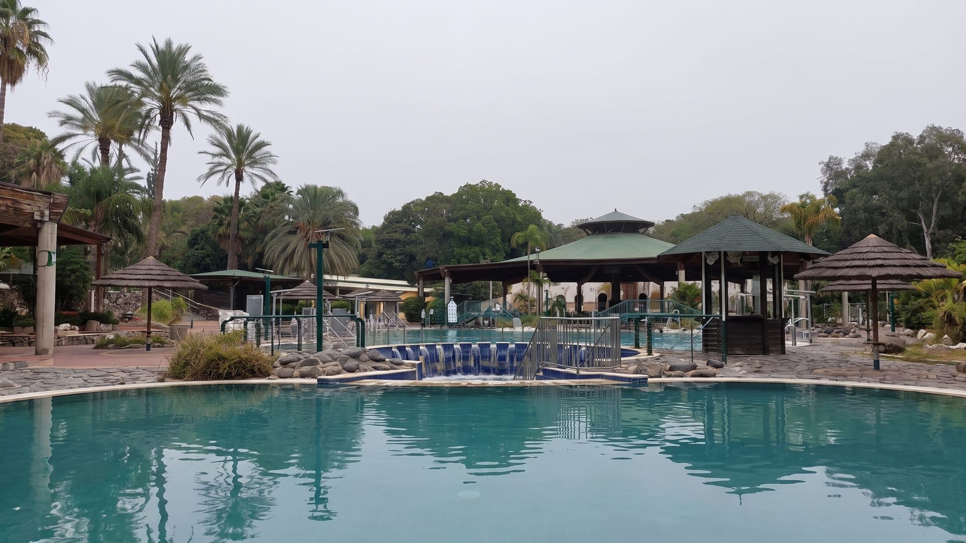 The thermal pool at Hamat Gader seen from the water's edge, with thatched umbrellas and palms along the far side