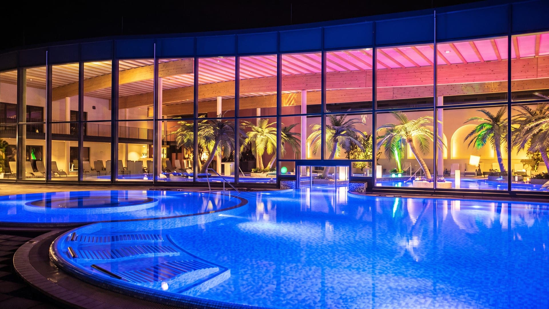 Palm-lined pool hall lit violet at night behind a wall of glass