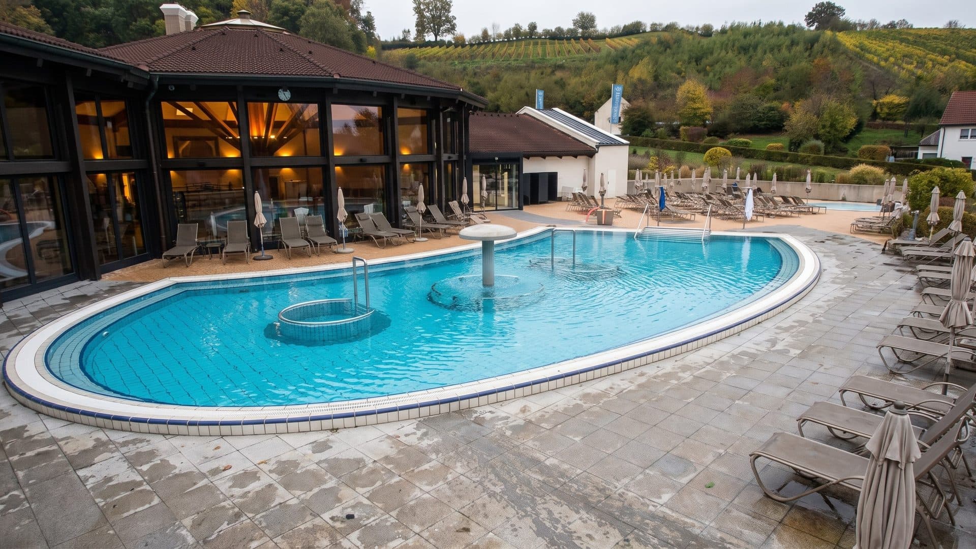 Oval outdoor thermal pool on a paved terrace with loungers, beside the timber-framed bath house and vineyard hills